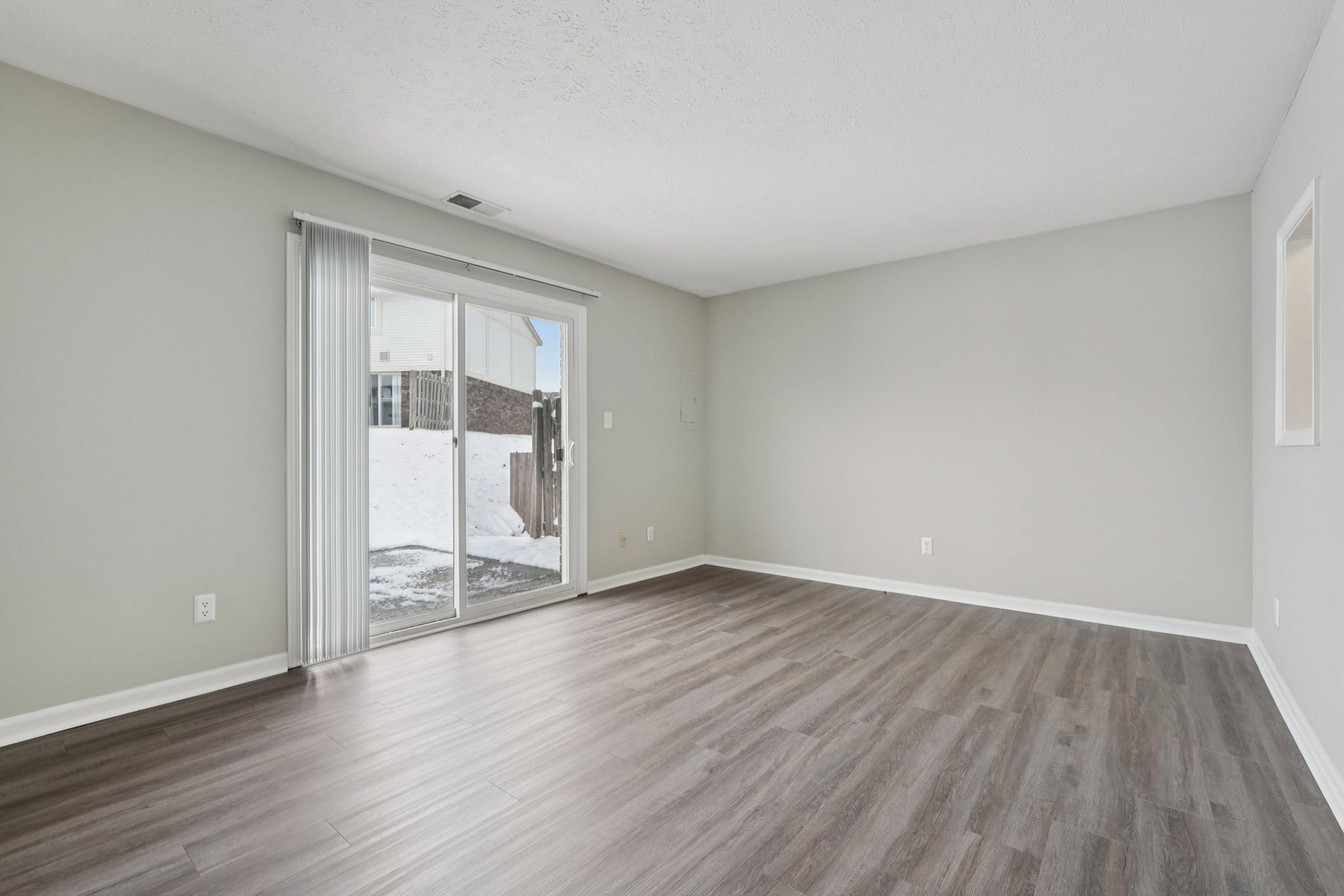 A spacious, empty room with light gray walls and dark laminate flooring. There are sliding glass doors leading to an outdoor area on one side, and a small window on the opposite wall. The area is well-lit with natural light coming from the doors.