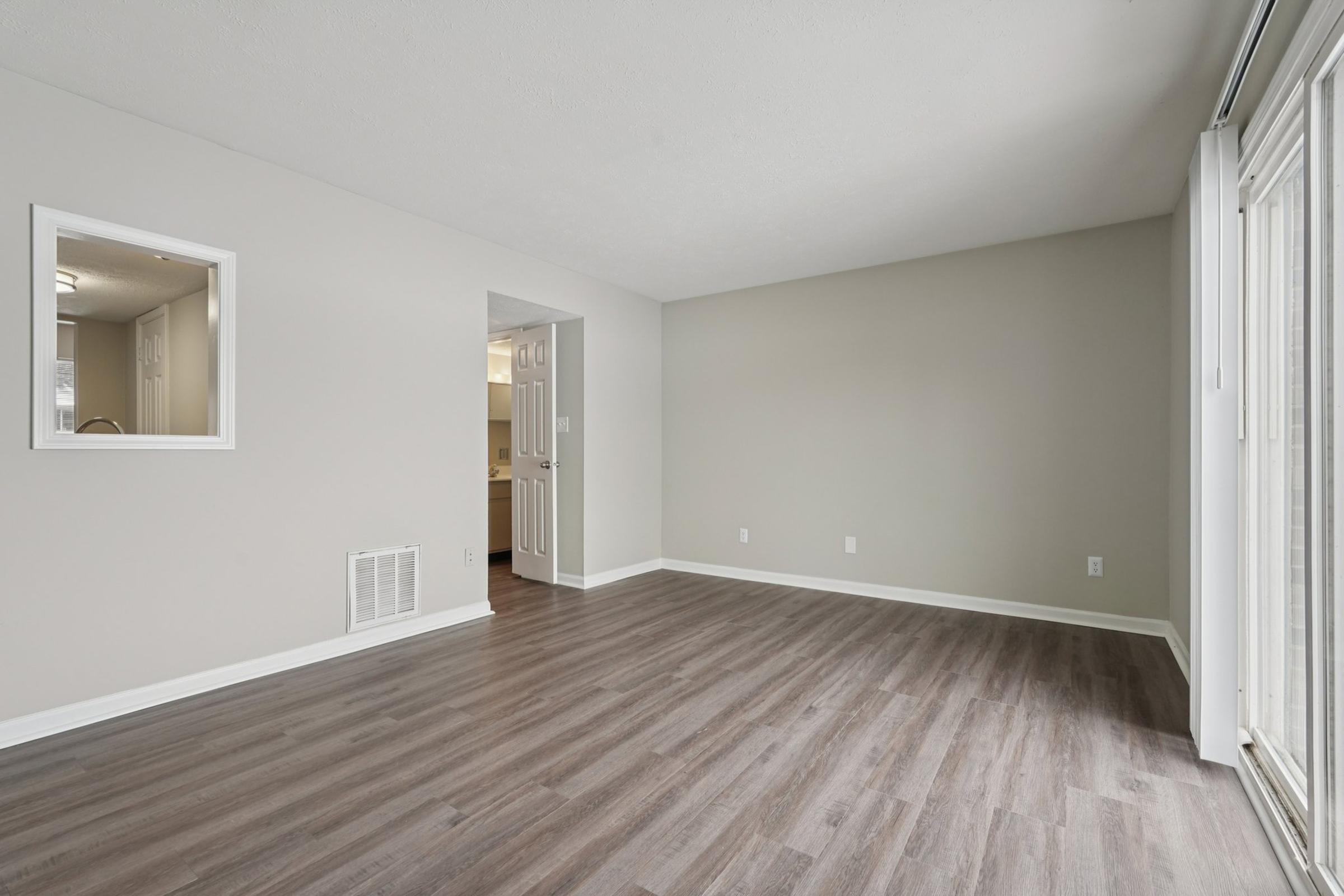 A spacious, empty living room with light gray walls and laminate flooring. A window with a small frame is visible on the left, and a doorway leads to another room on the right. The area is well-lit and features minimal decor, creating an open and airy atmosphere.