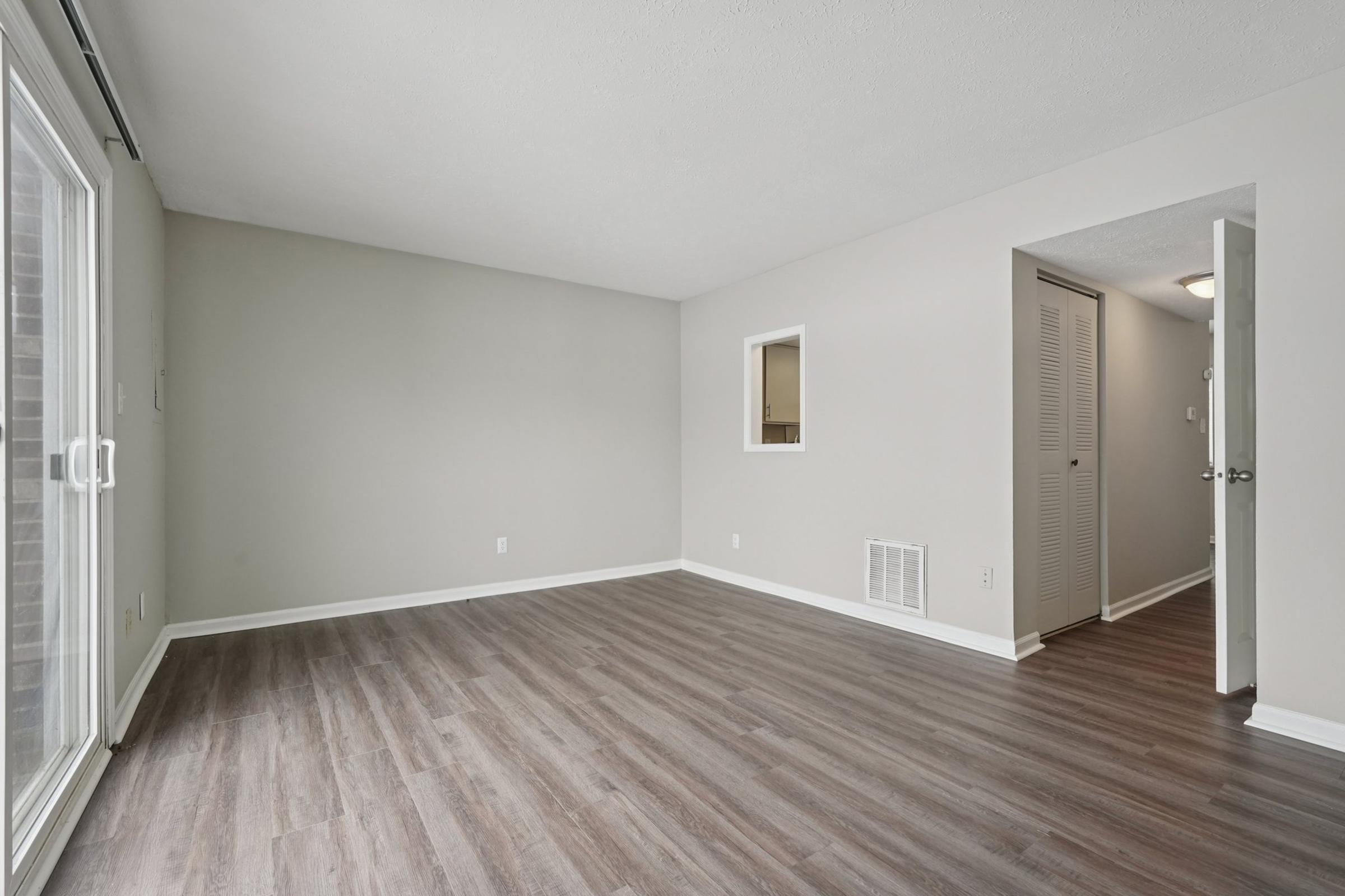 A spacious, empty living room featuring light gray walls and wood-style flooring. A large sliding glass door lets in natural light, while a small window and an entrance to another room are visible. The overall ambiance is bright and airy, suitable for personal decoration.