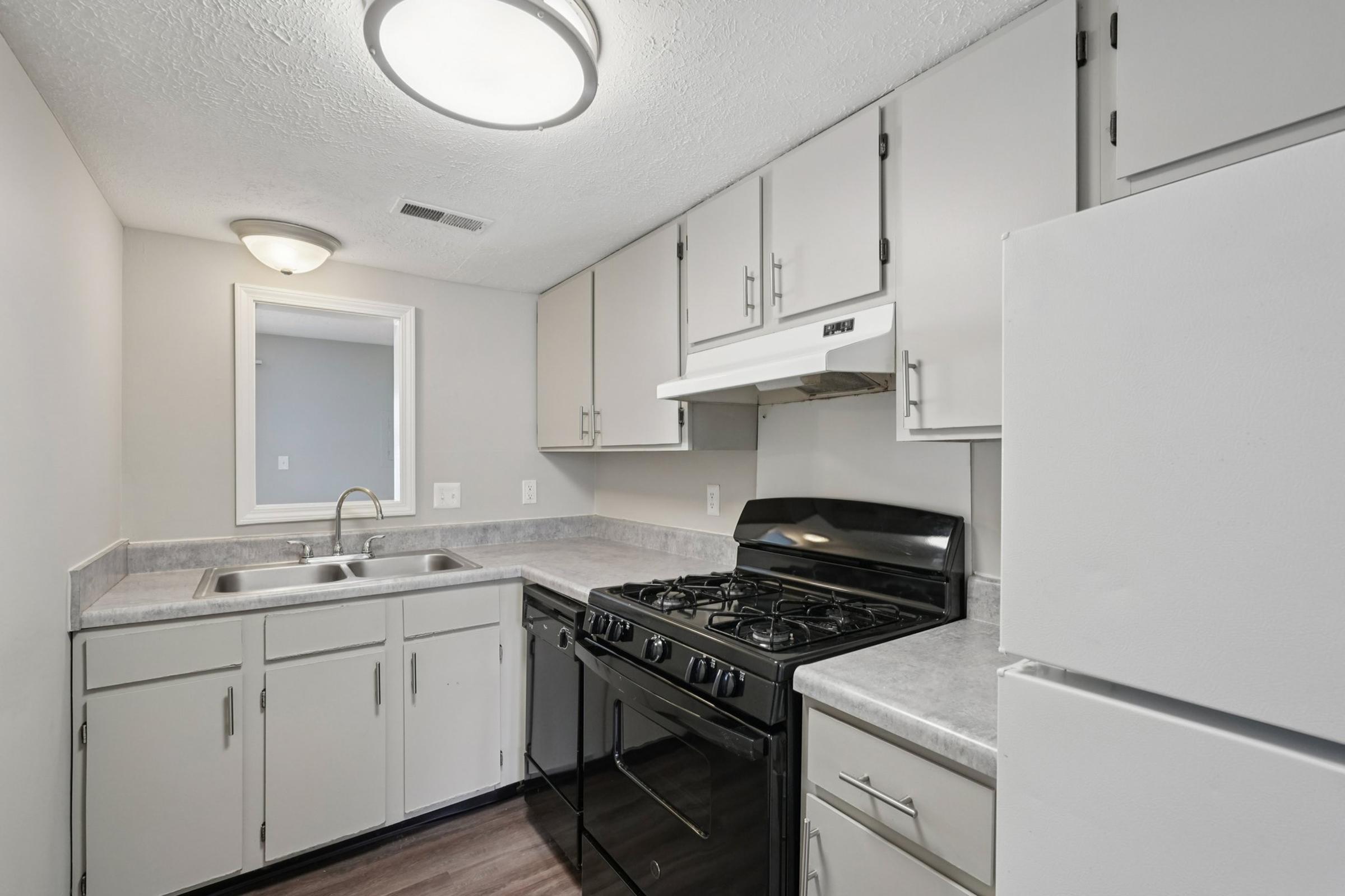 A modern kitchen featuring light-colored cabinets, a sink, and a gas stove. The countertop is grey, and there is a white refrigerator. The space has neutral wall colors and a ceiling light, creating a clean and functional appearance. A doorway leads to another room in the background.