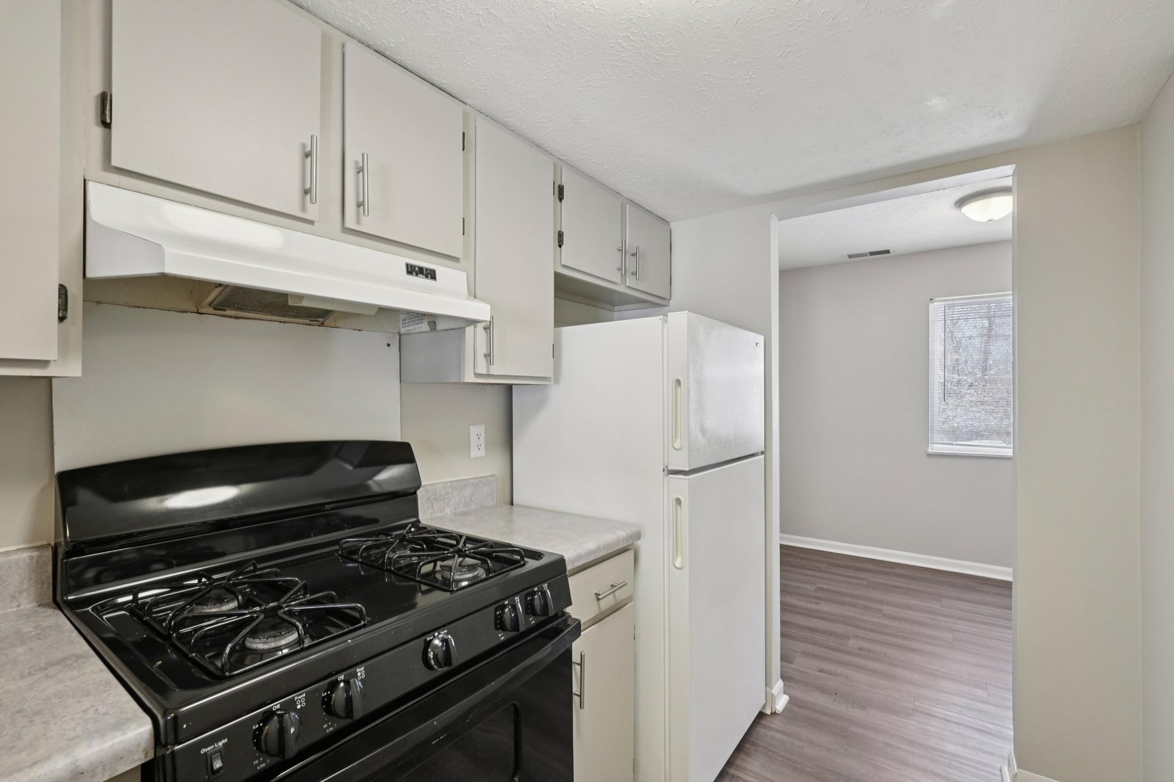 A kitchen with light gray cabinets, a black gas stove, and a white refrigerator. Countertops are gray. There's a window on the right-side wall leading to a room with hardwood flooring and neutral-colored walls. The overall aesthetic is modern and functional.