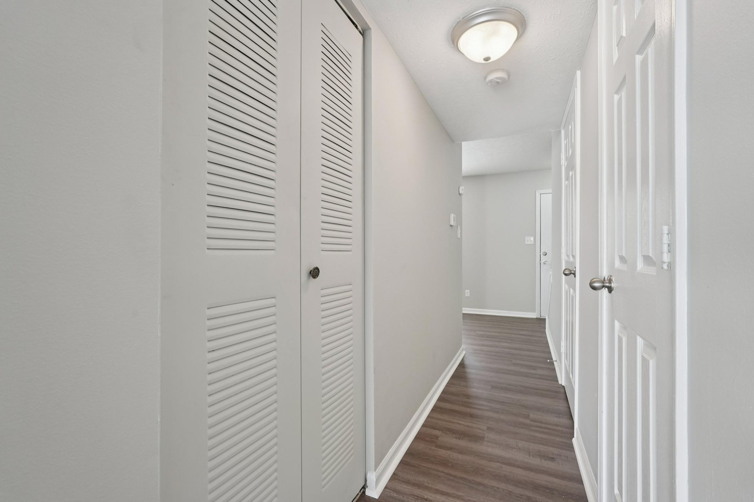 A well-lit hallway featuring closed double doors with louvered design on the left, leading to a light-colored wall. The floor is comprised of dark wood or laminate, and there are white doors leading to adjacent rooms. A ceiling light fixture illuminates the space, giving it a clean and modern appearance.