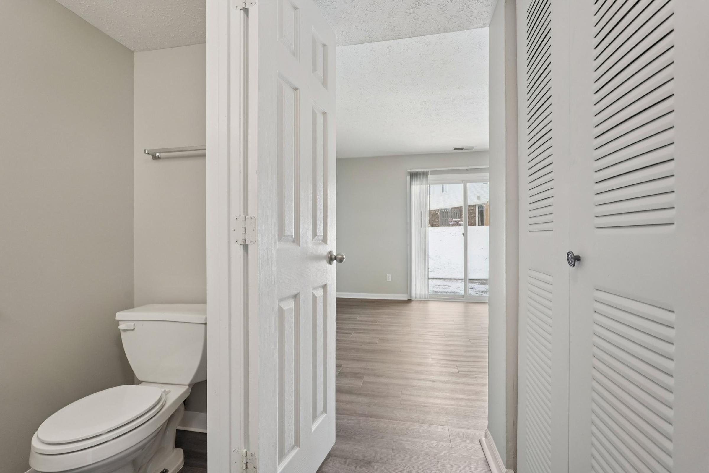 Interior view of a bathroom doorway, showing a toilet and a hallway leading to a living space. The living area features large windows with natural light, and a glimpse of snow outside. The room has light gray walls and wooden flooring, with a closet on the right adorned with louvered doors.