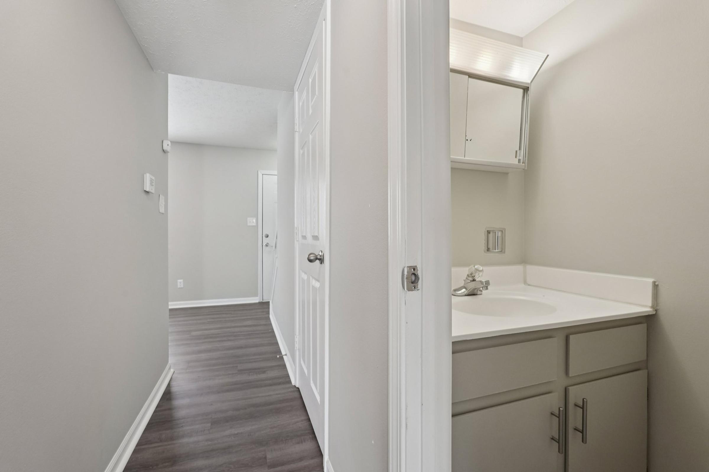 A narrow hallway leading to a door, with a small bathroom on the left. The bathroom features a sink and mirror with minimal decor. The walls are light gray, and the flooring is dark, wood-like laminate. The space is well-lit, creating a clean and modern atmosphere.