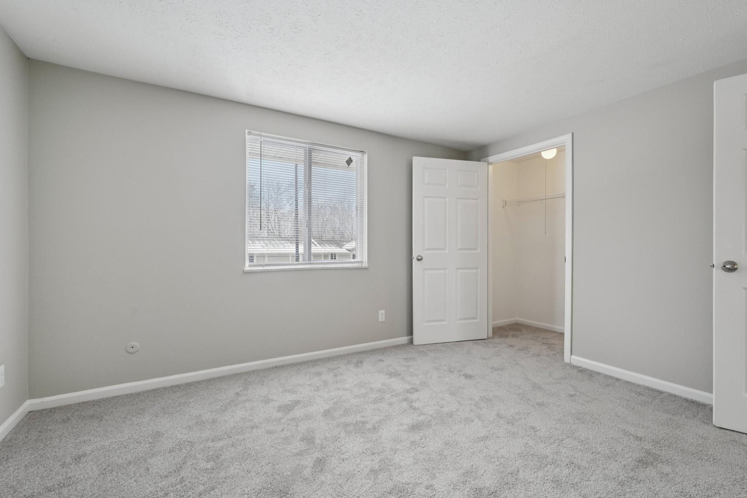 A bright, empty bedroom with light gray walls and new carpet. A window allows natural light to fill the space, and a door leads to a closet with a hanging rod. There is minimal furniture and decor, creating a clean and spacious atmosphere.