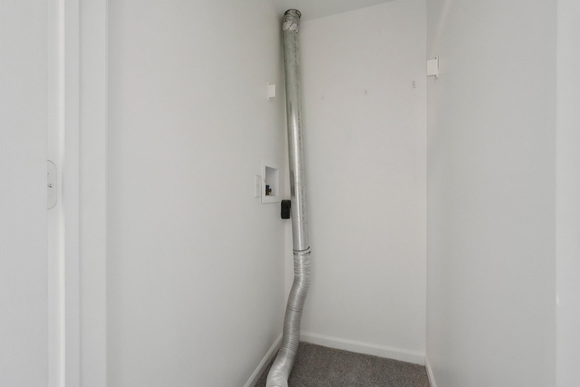 Narrow hallway with white walls and carpeting, featuring a metallic duct running vertically along one side. A small opening is visible on the left wall, and there are two wall hooks mounted above. The space is simple and uncluttered.