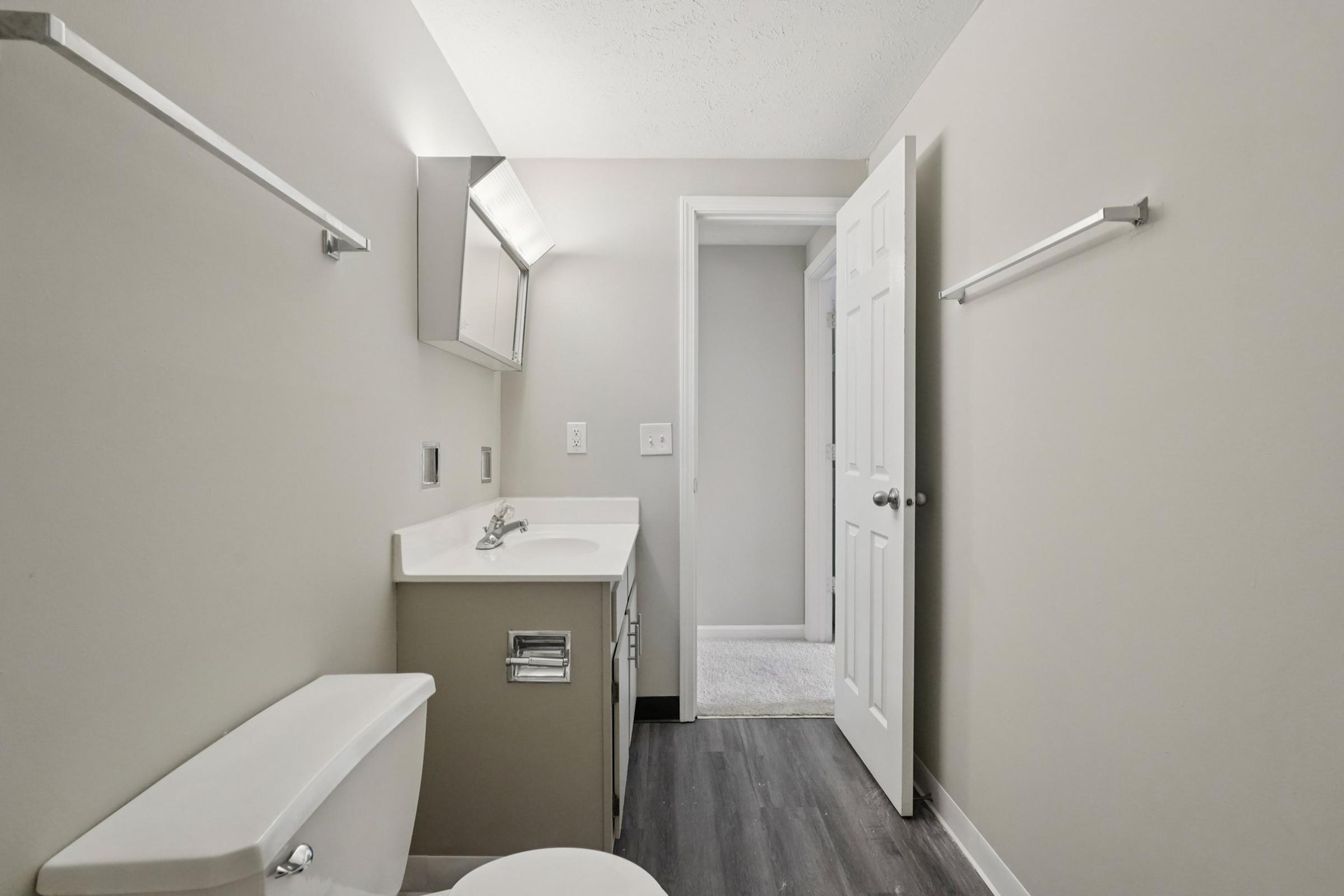 A small, modern bathroom featuring a light gray color scheme. It has a white toilet, a vanity with a sink, and a large mirror. Two towel bars are on the wall, and a door leads to an adjacent room. The flooring appears dark and contrasts with the lighter walls and fixtures.