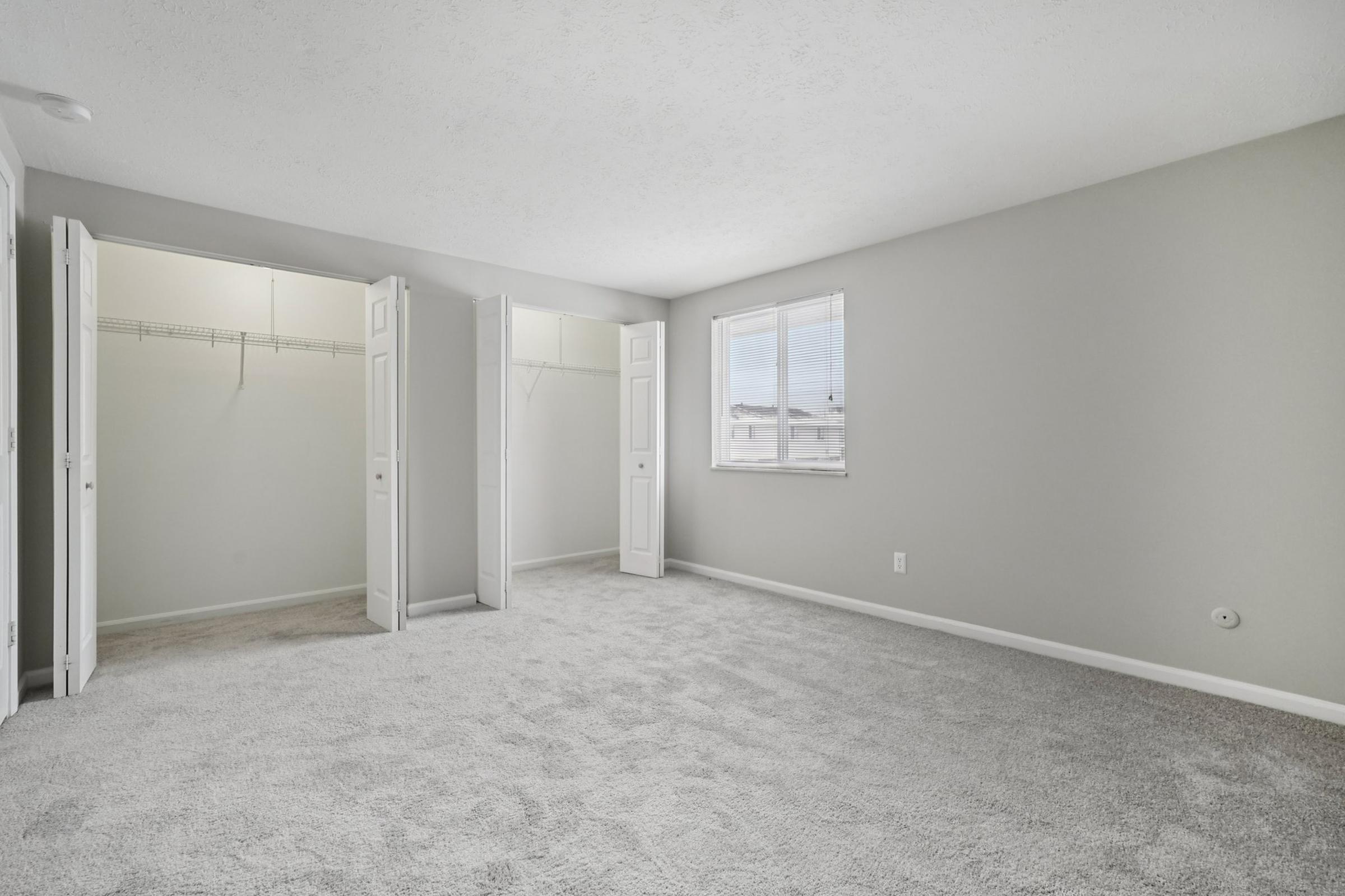 A spacious bedroom with light gray walls and soft carpet flooring. The room features two mirrored closet doors on one side and a window allowing natural light to illuminate the space. The overall atmosphere is clean and inviting, perfect for personalizing with furniture and decor.