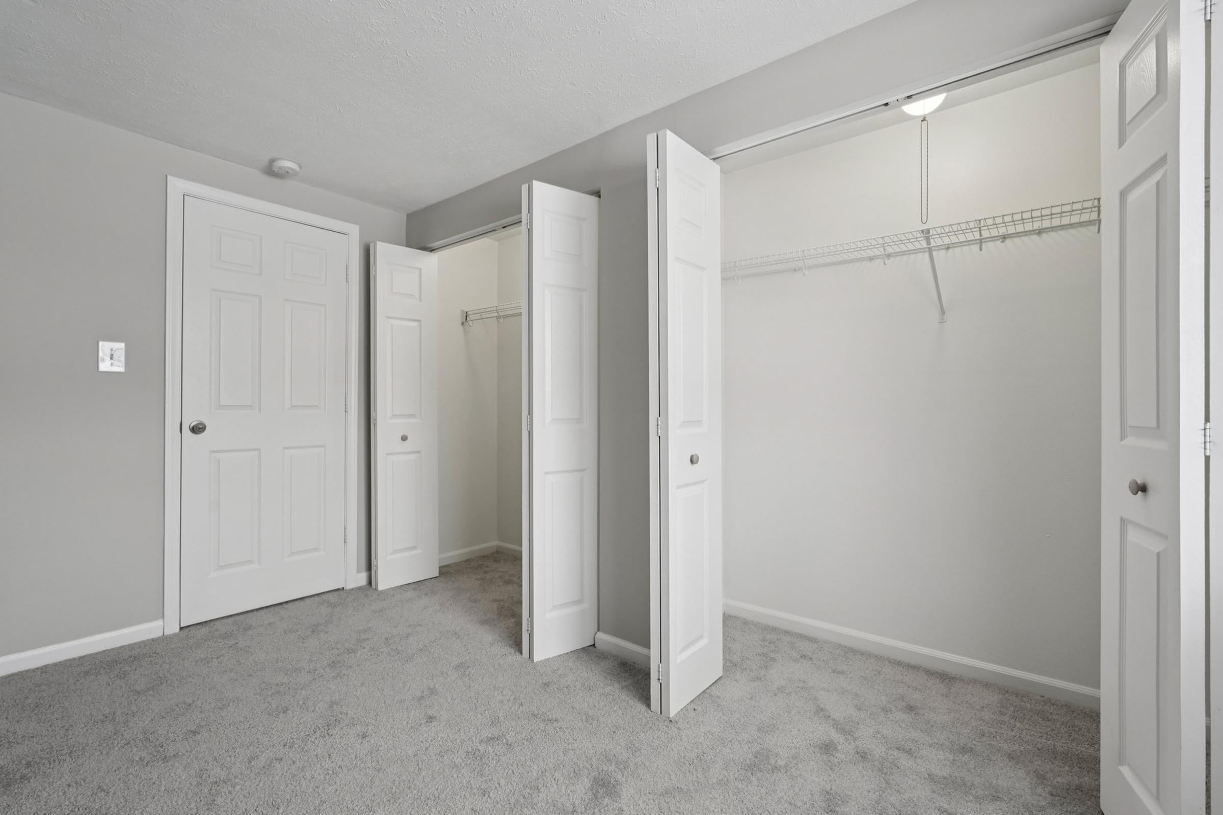 A well-lit, empty bedroom featuring light gray walls and plush gray carpeting. Two open double doors lead to a spacious closet with wire shelving. The closet area is bright, showcasing the clean and minimalistic design of the room.