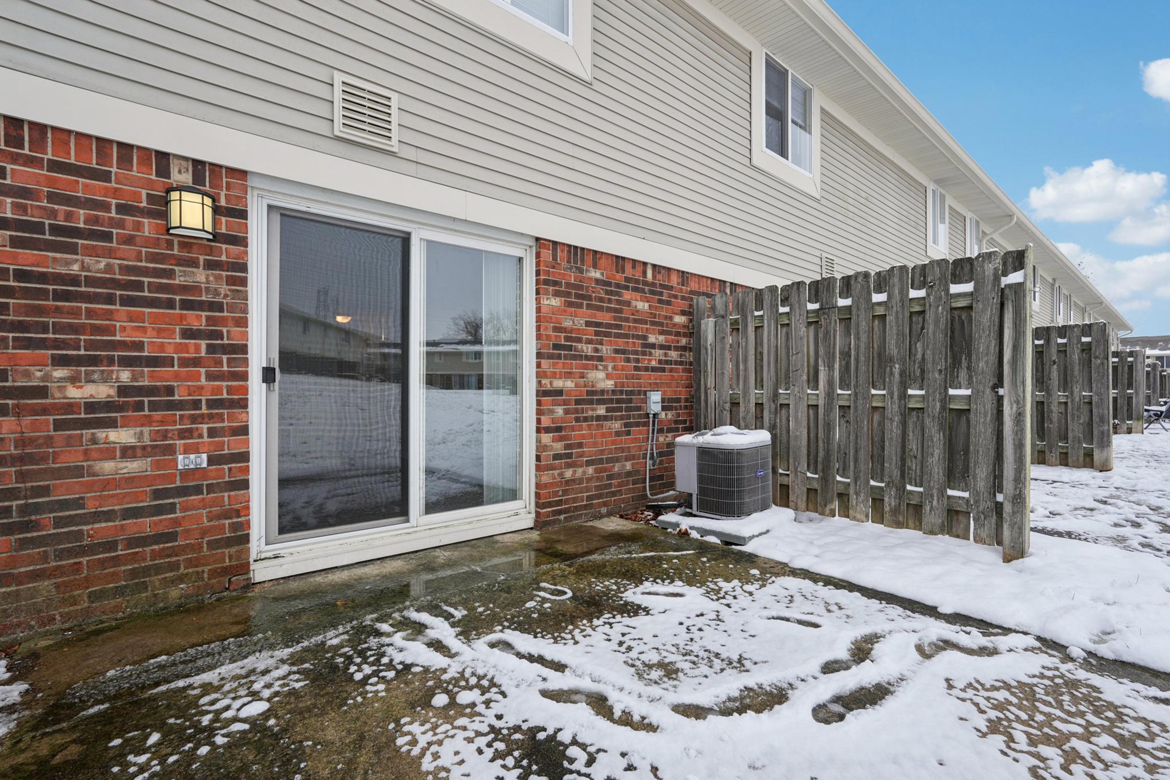 A view of a patio area behind a house, featuring a sliding glass door, a wooden fence, and a snow-covered concrete surface. An outdoor air conditioning unit is visible next to the door, and the surrounding area shows a light dusting of snow. The sky is clear with a few clouds.