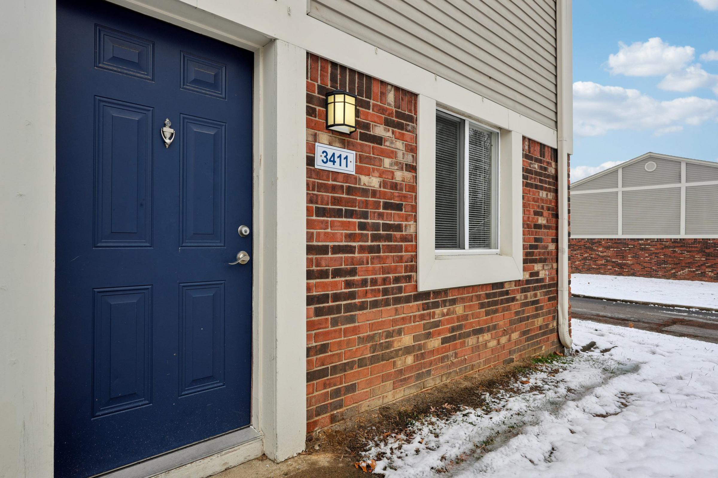 A corner view of a building entrance featuring a blue front door with a doorbell and a decorative light fixture. The adjacent wall is made of red brick, and there is a small patch of snow on the ground. A visible address number, 3411, is placed near the door. The sky is partly cloudy.