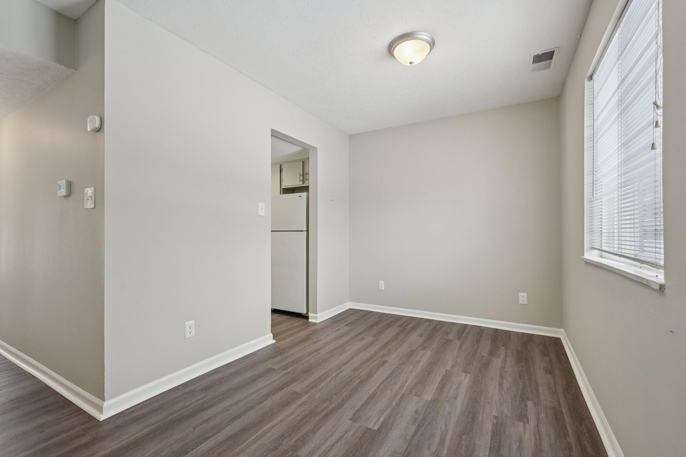 Empty room with light-colored walls and a wooden floor. A ceiling light fixture is visible, along with a small window letting in natural light. A doorway leads to a kitchen area, which has a white refrigerator partially visible. The space appears clean and well-maintained, suitable for various uses.