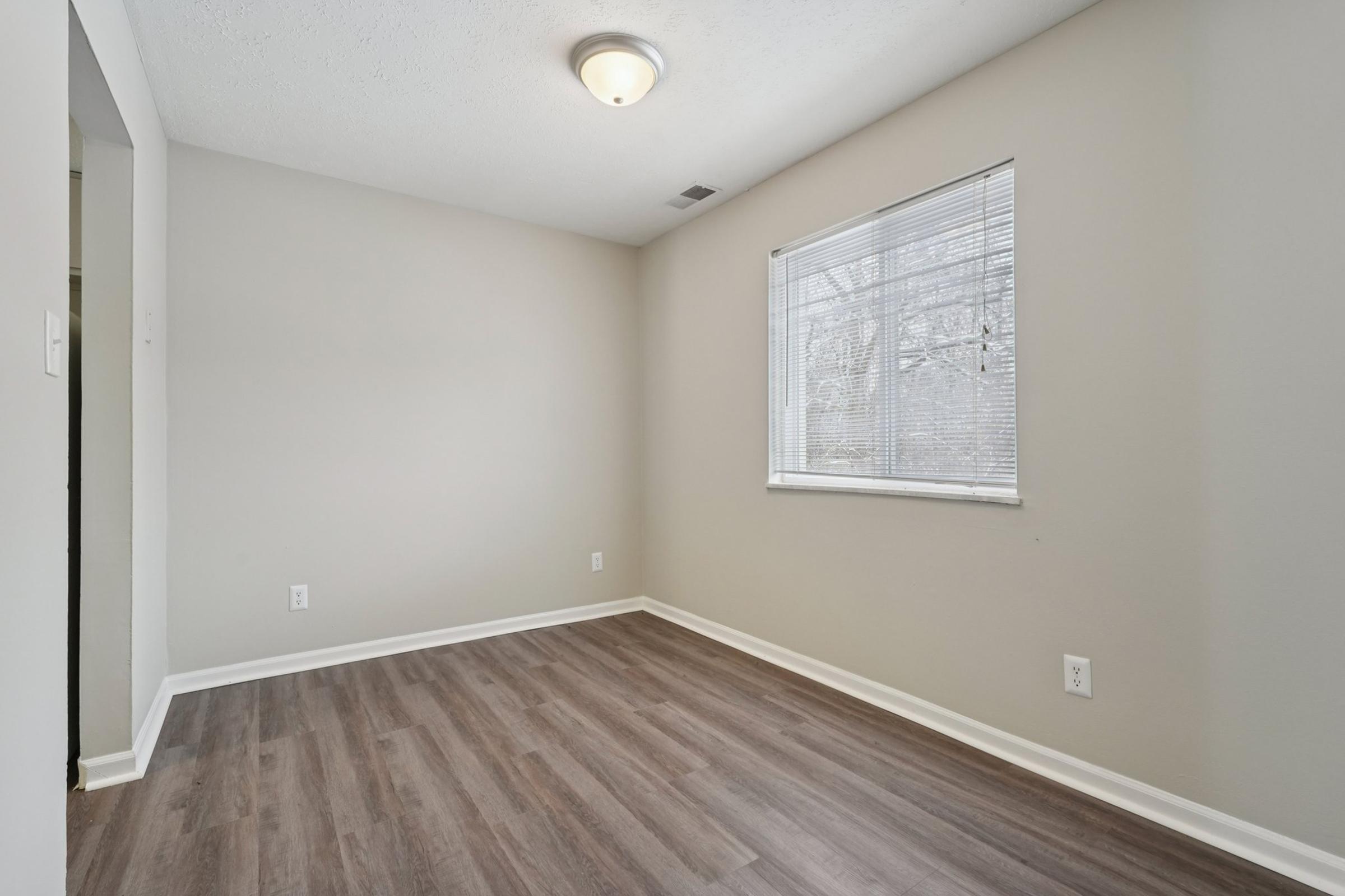 A small, empty room with light gray walls and a window letting in natural light. The floor is covered with wood-like laminate, and there is a ceiling light fixture. The room is unfurnished, creating a spacious and minimalist atmosphere.
