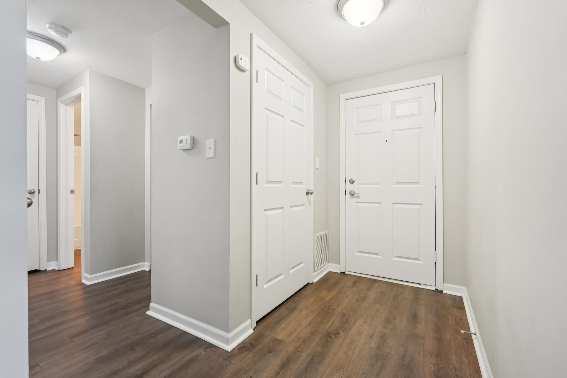 Interior view of a well-lit hallway featuring a front door with a window, two other closed doors on the left, and light-colored walls. The floor is covered with laminate wood, and there are ceiling lights providing illumination. The space appears clean and modern.