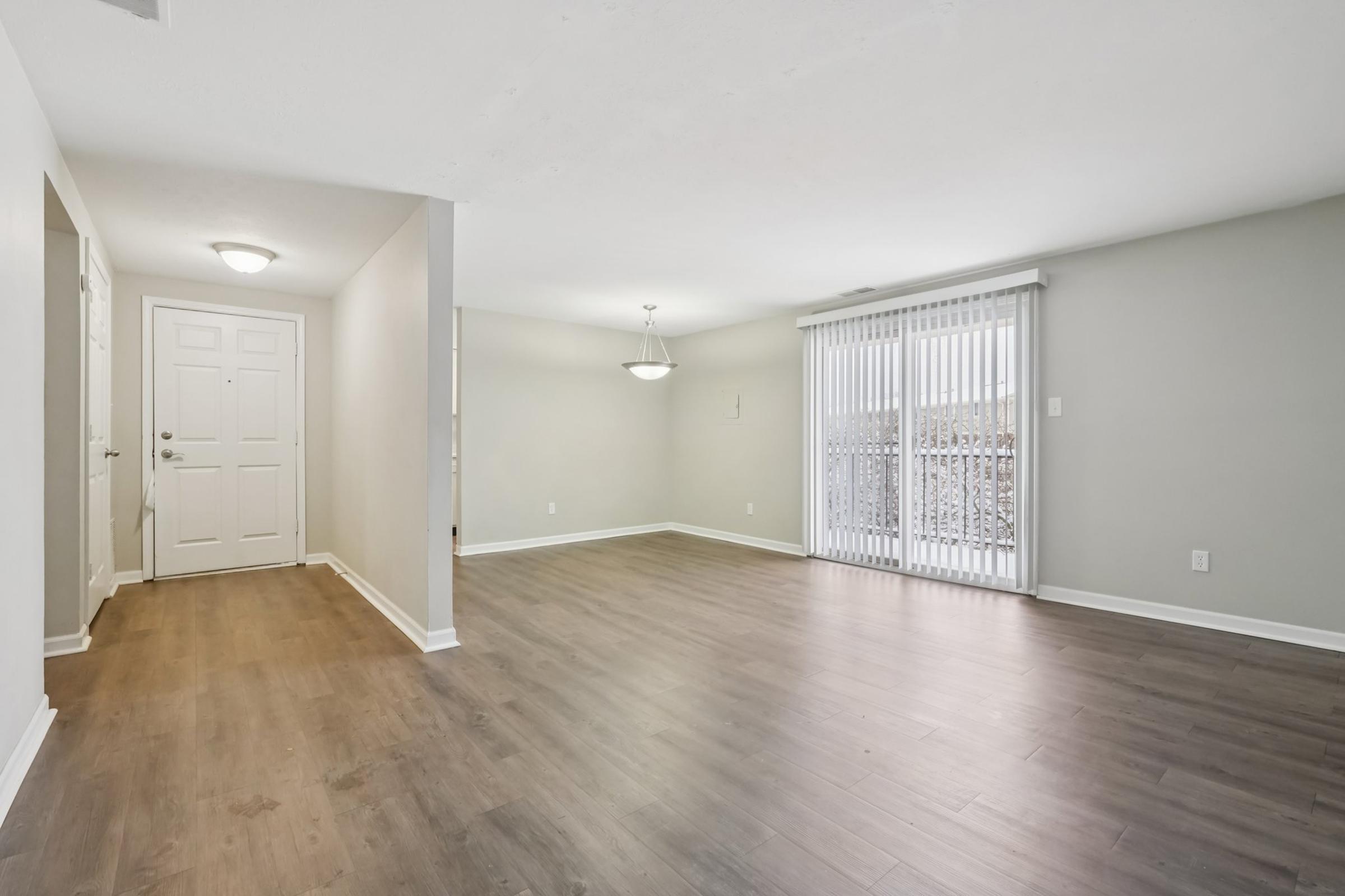 Spacious, empty living room with light-colored walls and wooden flooring, featuring a door leading outside, a single pendant light, and large windows with vertical blinds. The area is well-lit and has a neutral, modern aesthetic, ideal for personalization.