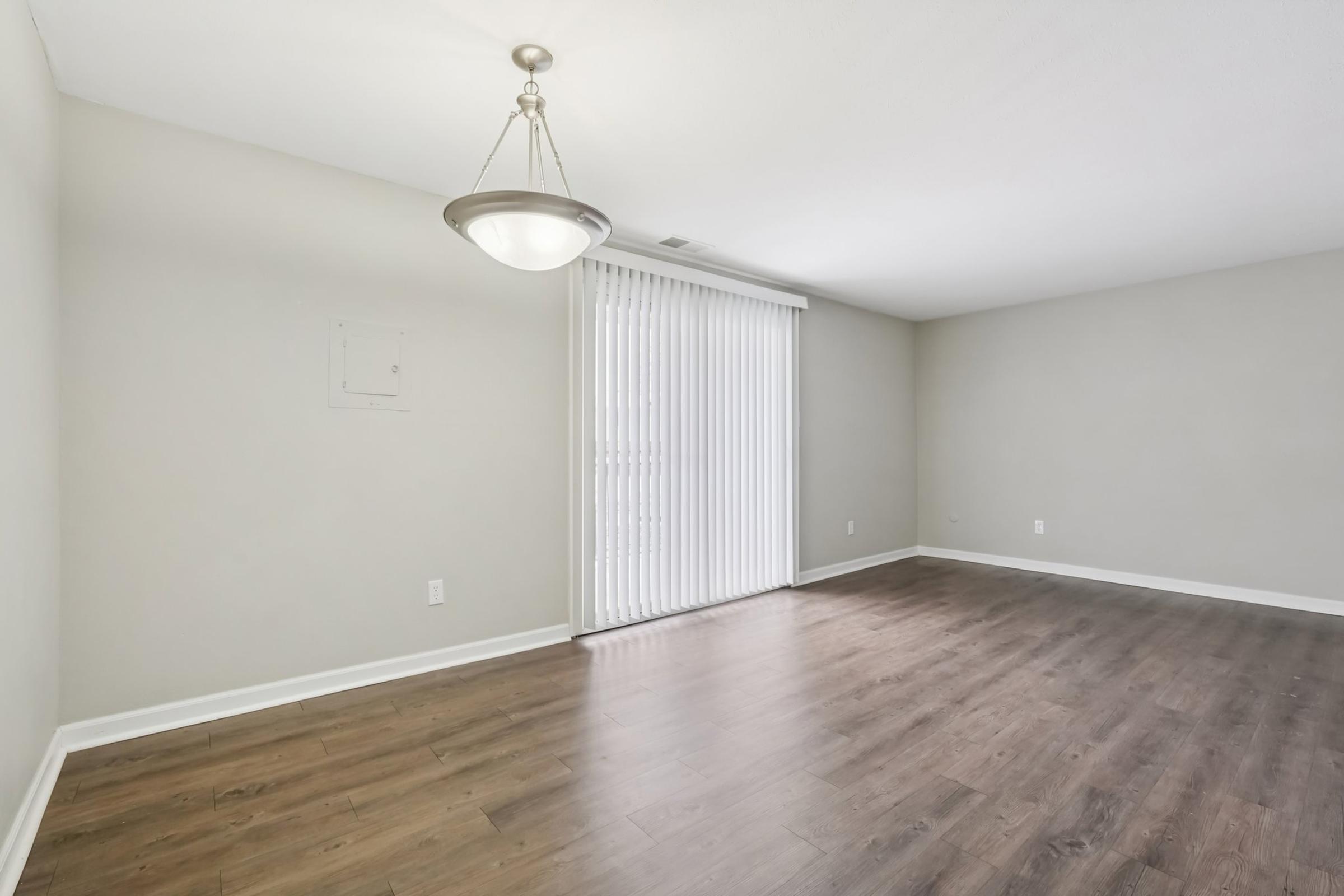 A spacious, empty room with light-colored walls and a wood laminate floor. A ceiling light fixture hangs in the center, and large vertical blinds cover a window on one side, allowing natural light to fill the space. The room is ready for decoration or furniture placement.