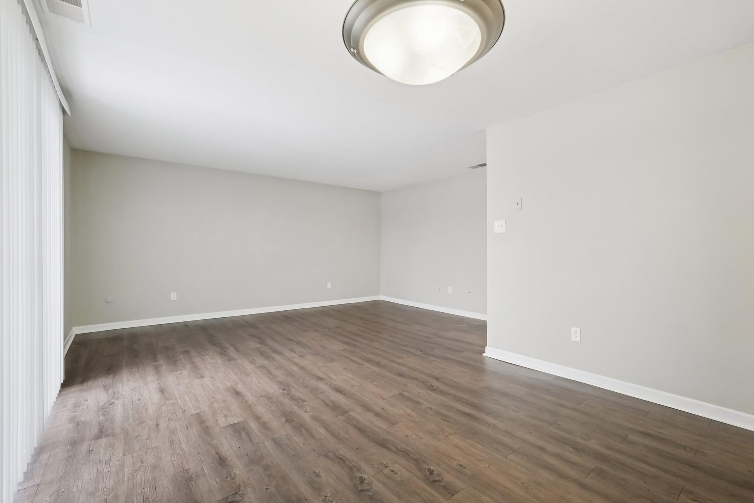 A spacious, empty room with light-colored walls and wood-like flooring. A ceiling light fixture is visible, and large windows with vertical blinds occupy one wall, allowing natural light to fill the space. The room is devoid of furniture, creating an open and airy atmosphere.