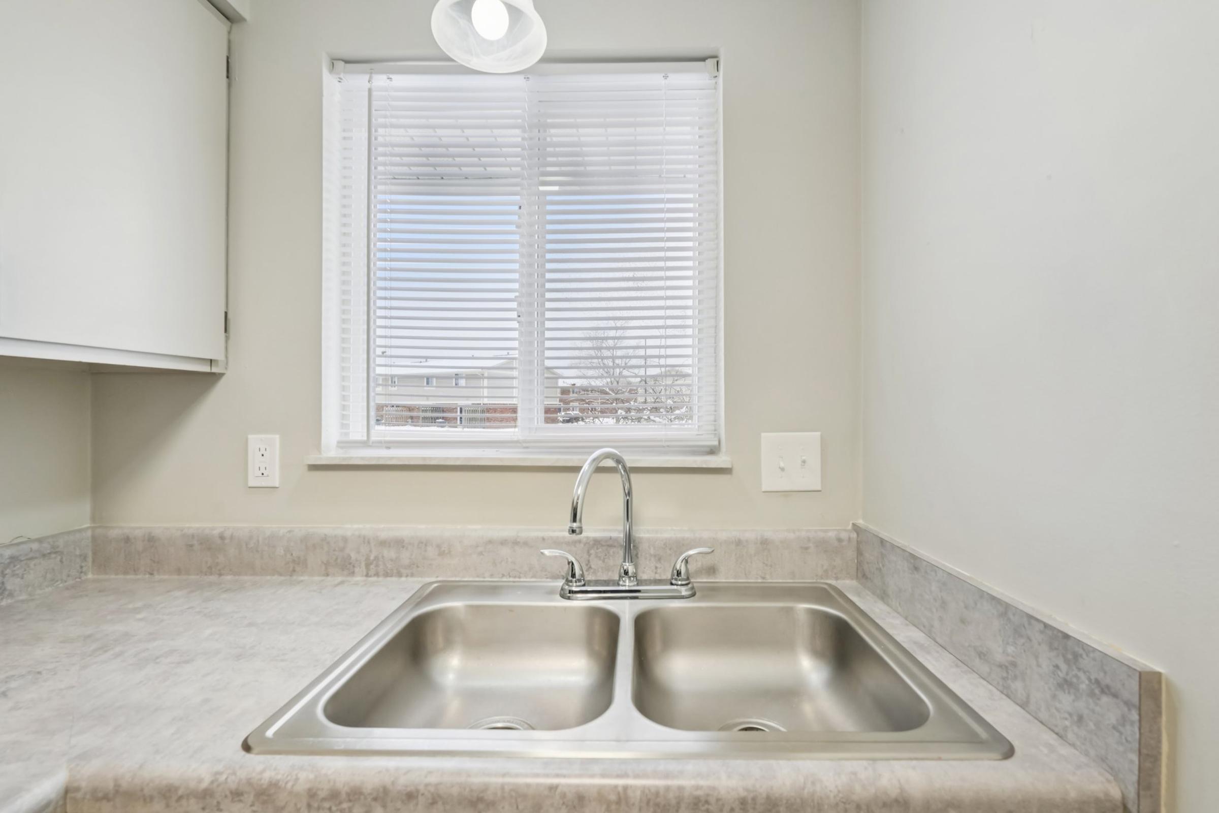 A modern kitchen sink with a double basin, set against a light-colored countertop. The window above the sink features white blinds, allowing natural light to enter the space. The overall room has a neutral color palette, contributing to a clean and functional kitchen design.