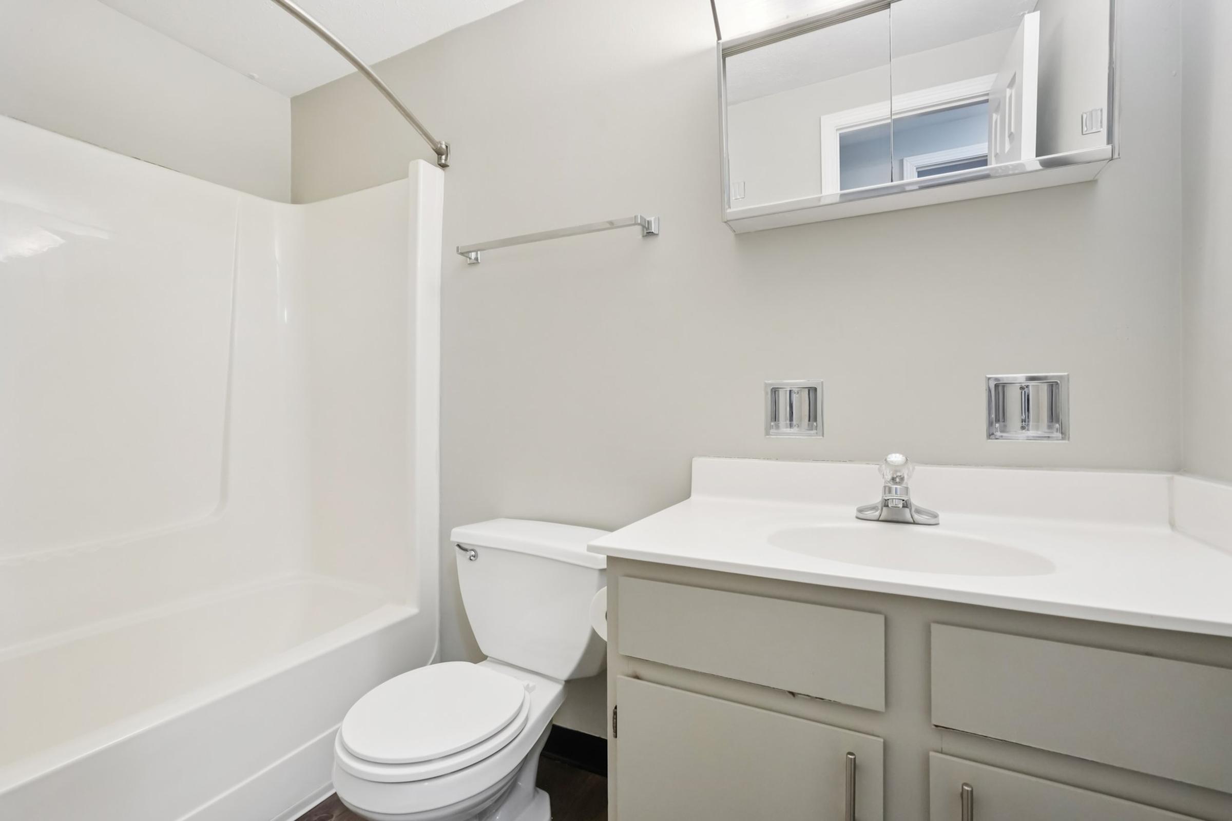 A clean and modern bathroom featuring a white bathtub with a shower, a toilet, and a sink with a countertop. The walls are painted in a light gray, and there is a mirror above the sink. The overall aesthetic is simple and uncluttered, highlighting functional bathroom elements.
