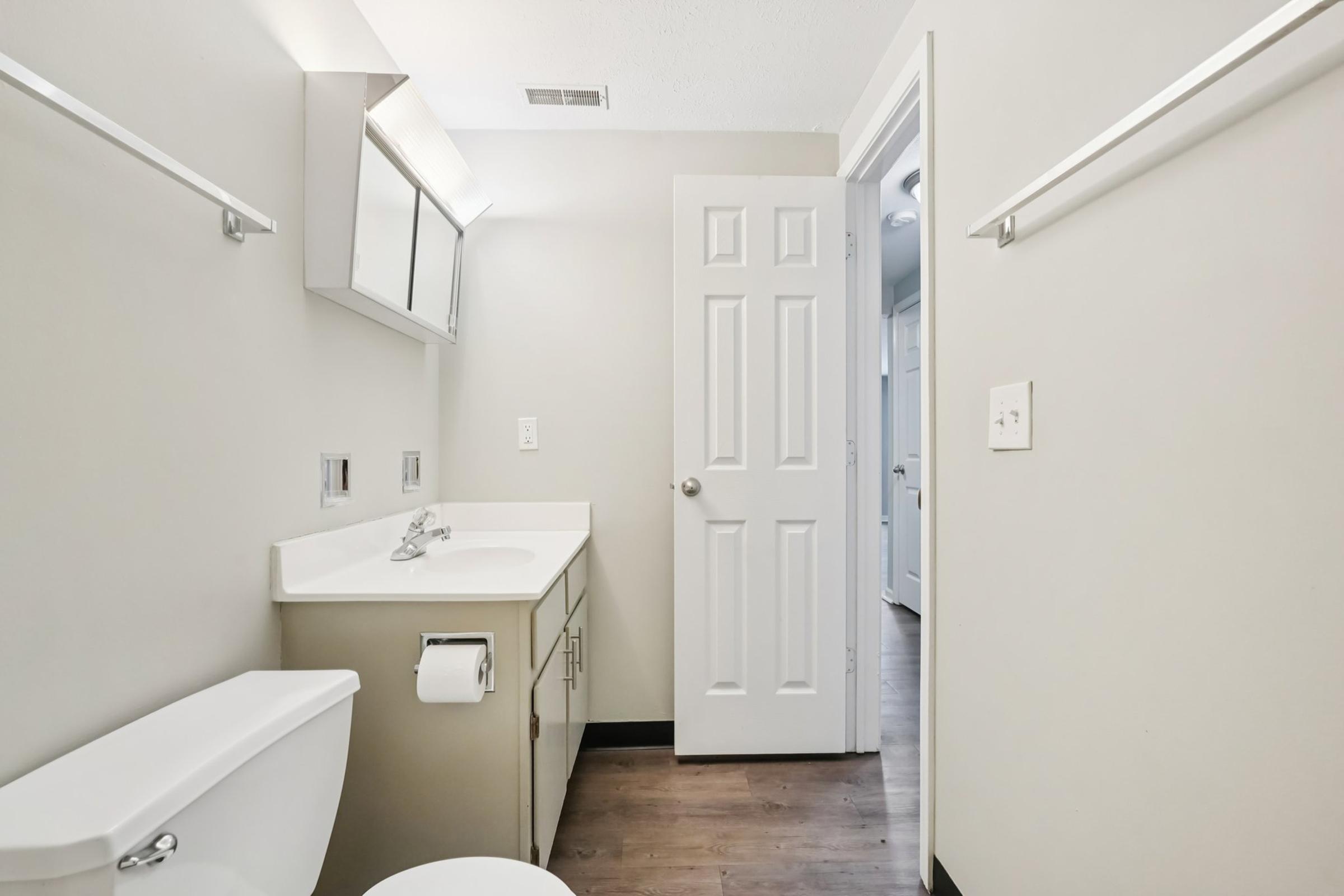 A clean, modern bathroom featuring a white sink and cabinet, a toilet, and a mirror above the sink. The walls are painted a light gray, and the floor has a dark wooden appearance. A door leads to another room, and there are minimal shelves on the walls.