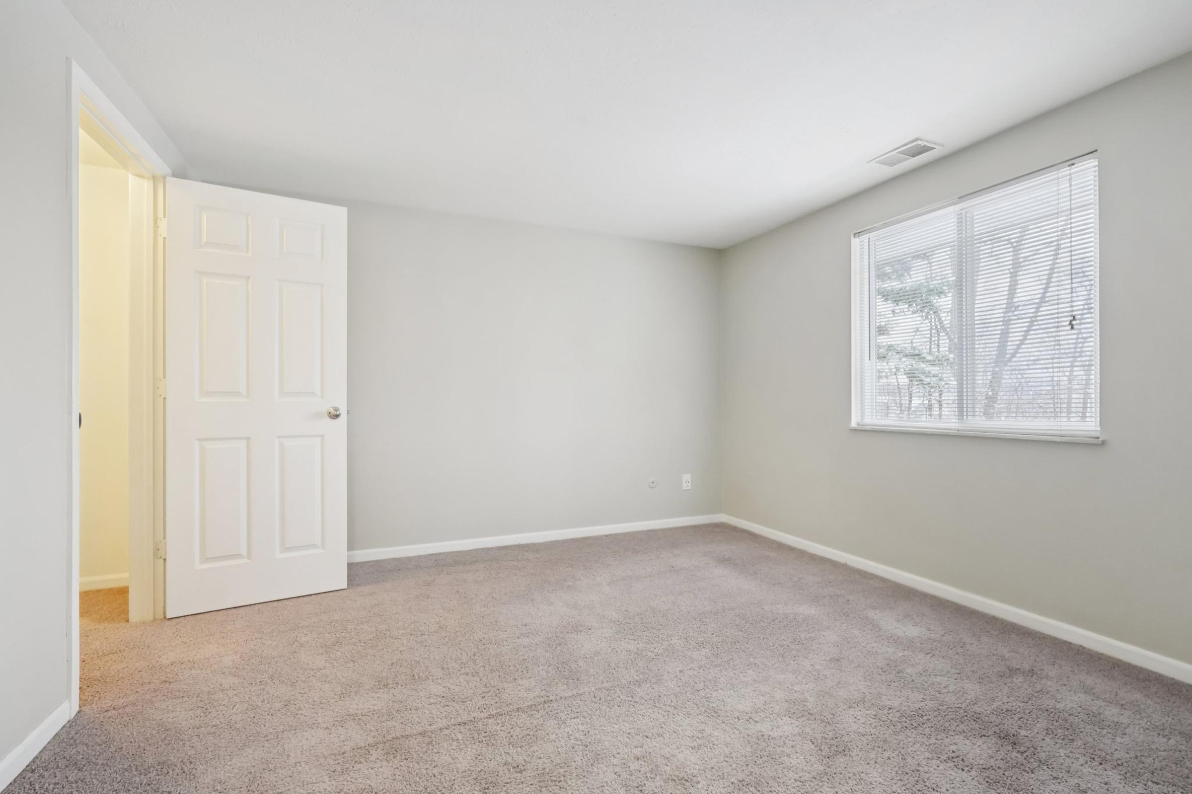 A light-filled, empty room with beige walls and new carpet. There's a closed white door on the left and a window with blinds on the right, providing a view of the outdoors. The space appears clean and ready for furniture or decoration, making it suitable for various uses.