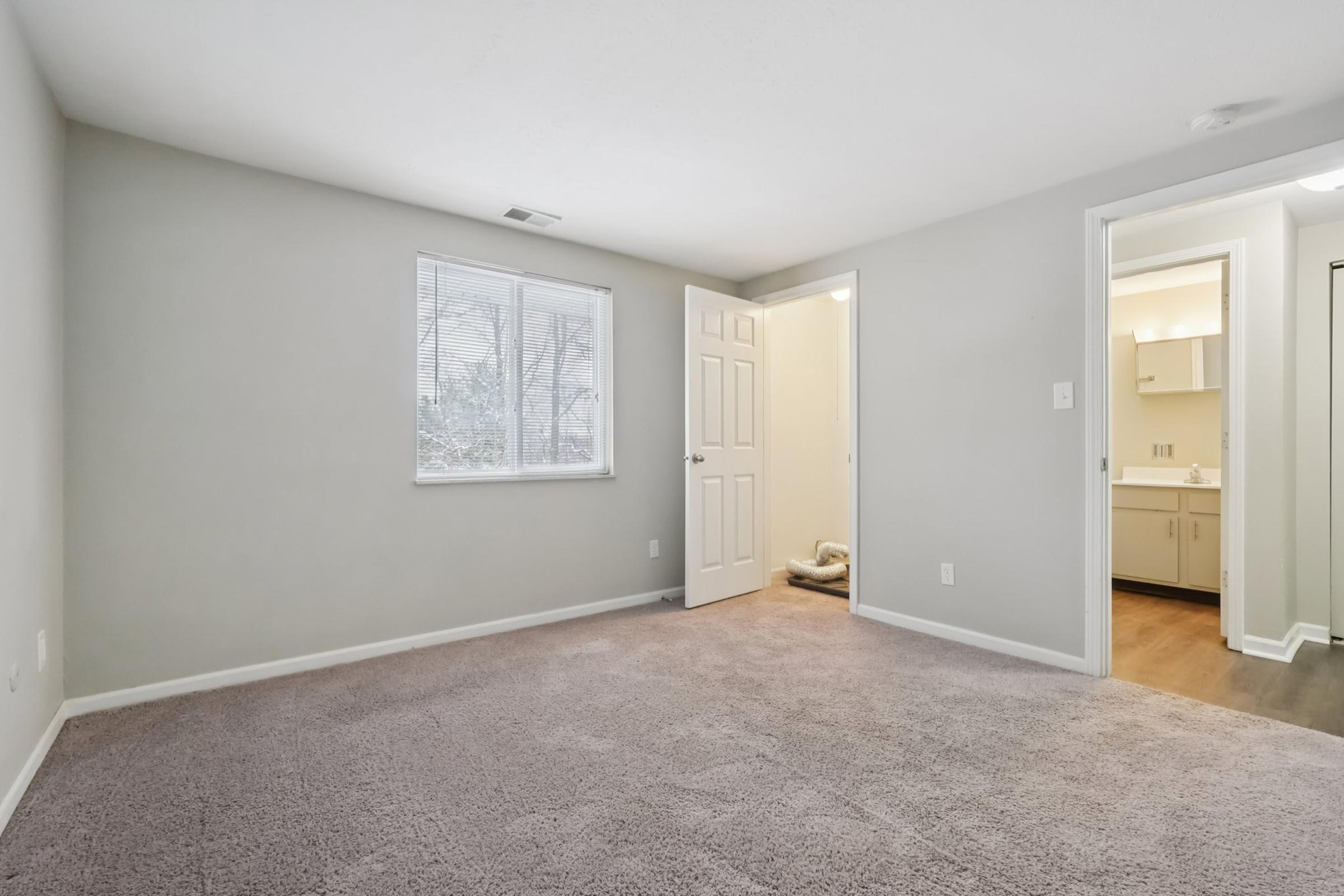 A spacious, neutral-toned room with light gray walls and plush carpet. There is a window allowing natural light, a closed door leading to another room, and a partially visible bathroom in the background. The overall atmosphere is bright and inviting, suitable for various uses.