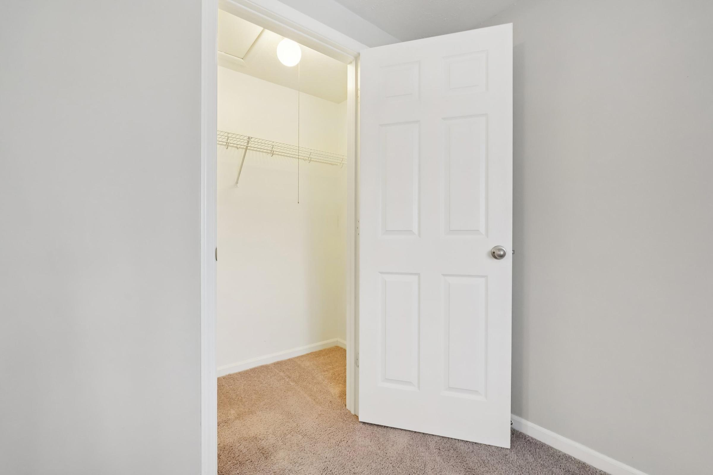 A view of a walk-in closet with a white double door slightly ajar. The interior has a bare wall with a wire shelving unit and a light fixture on the ceiling. The floor is carpeted in a neutral color, creating a clean and simple appearance.