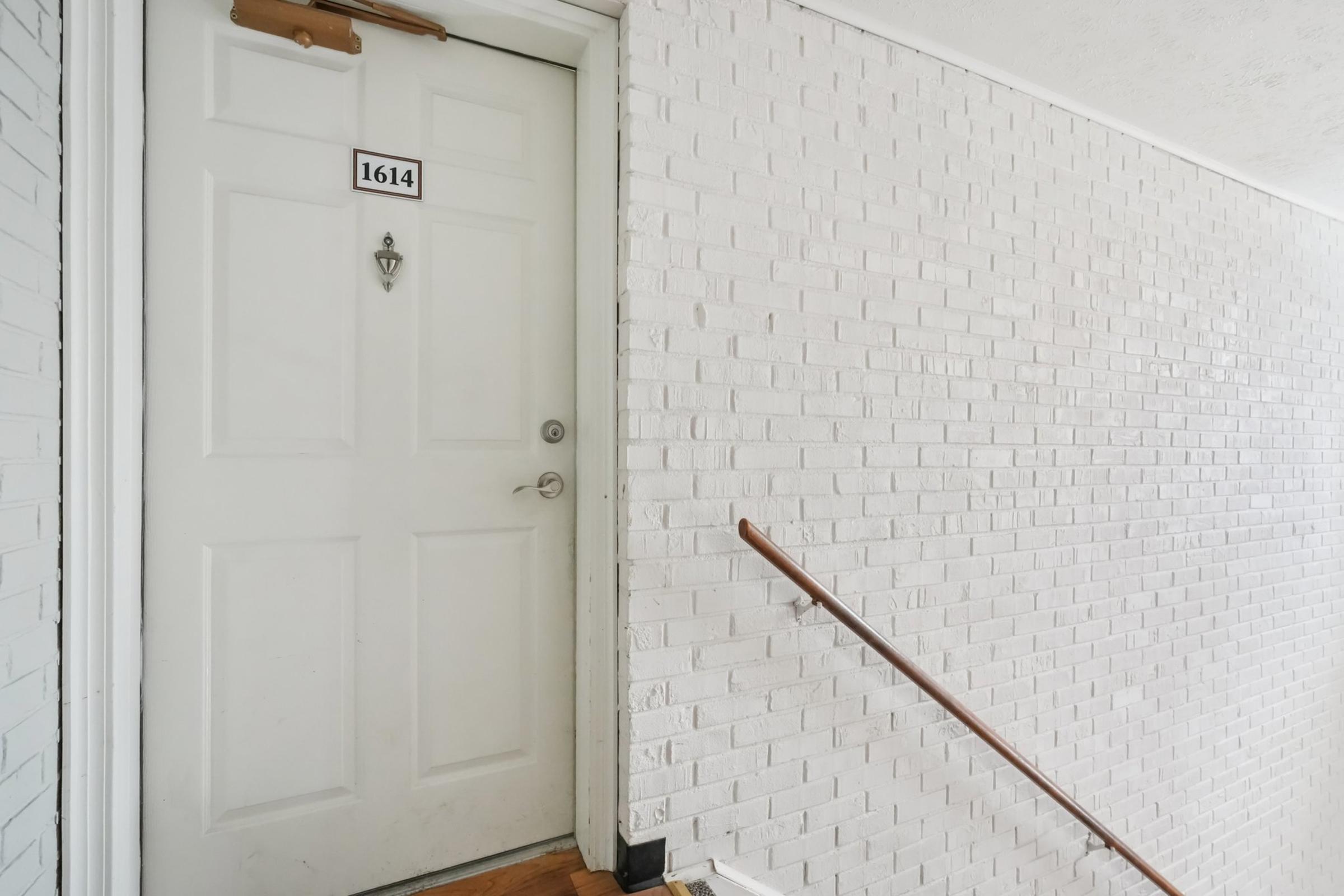 A white brick wall next to a door labeled "1614." The door has a small knocker and a doorknob. A wooden handrail runs along the wall next to a staircase that leads up to the door. The overall setting appears to be an indoor corridor or entryway.