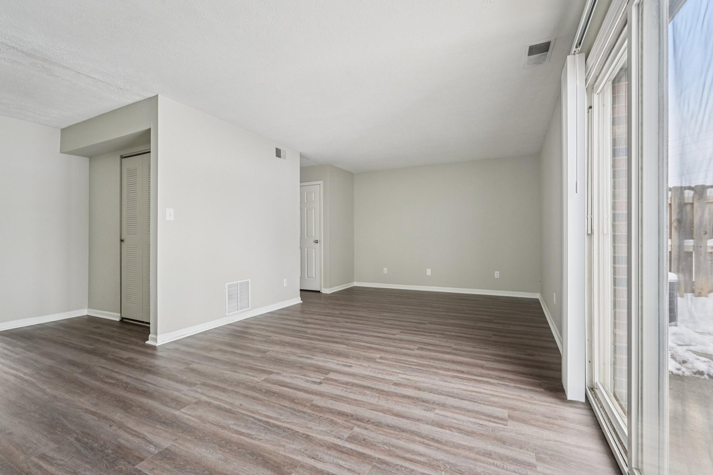 Spacious empty room with light gray walls and new laminate flooring. A large window allows natural light to fill the space, and a door leads to an adjoining area. The room is unfurnished, offering a blank canvas for potential decoration or use.