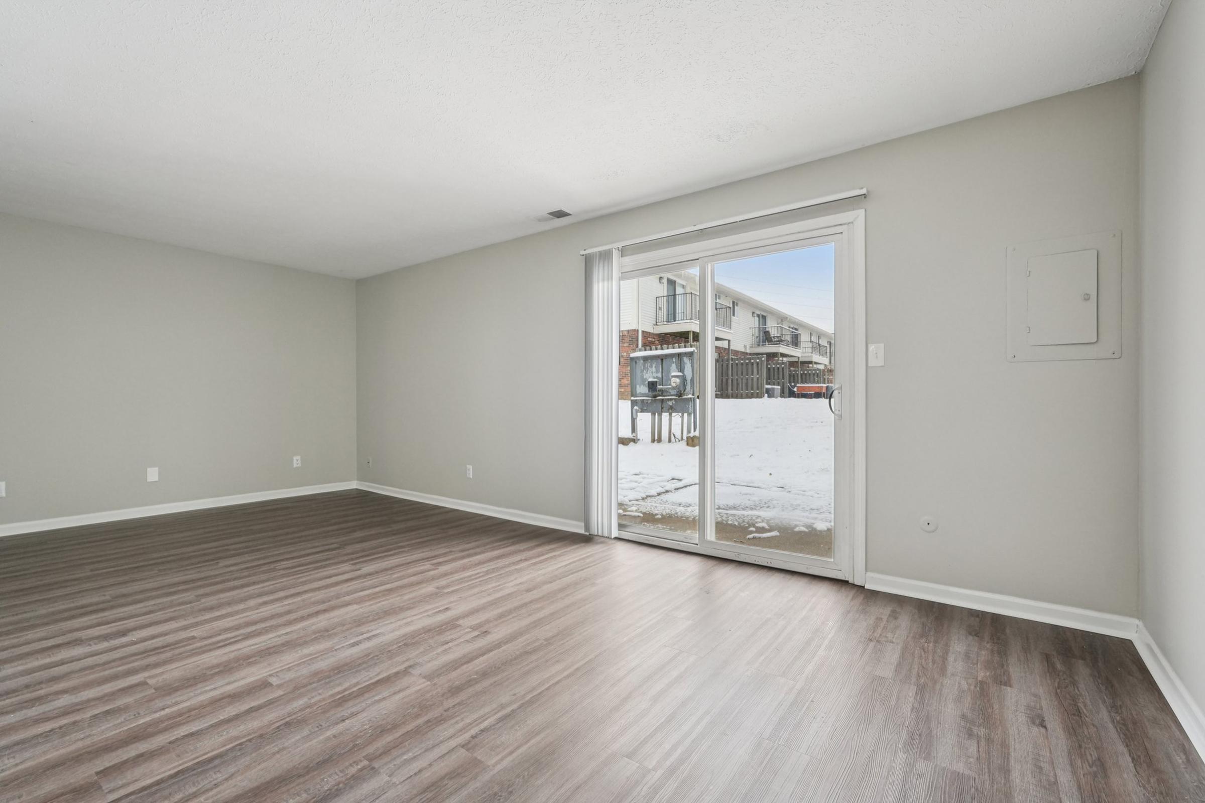 Spacious, empty room with light gray walls and new laminate flooring. A sliding glass door leads to an outdoor area with snow in the background. There is an electrical panel on one wall, and ample natural light is coming in from the outside.