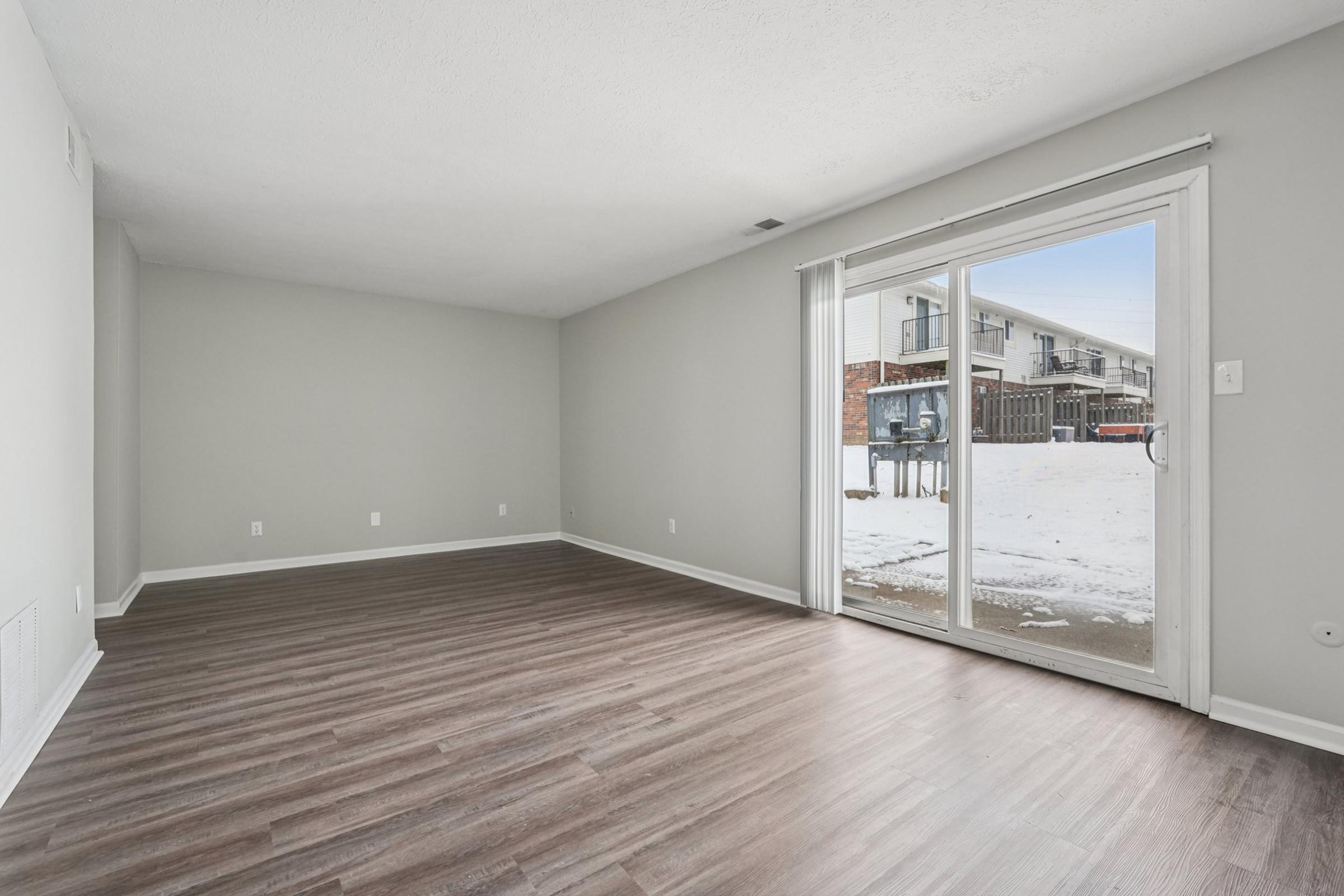 A spacious, empty living room featuring light gray walls and a large sliding glass door that opens to a snowy outdoor area. The floor is covered with modern laminate flooring, and there are no furnishings or decorations in the room, creating a fresh and inviting ambiance.