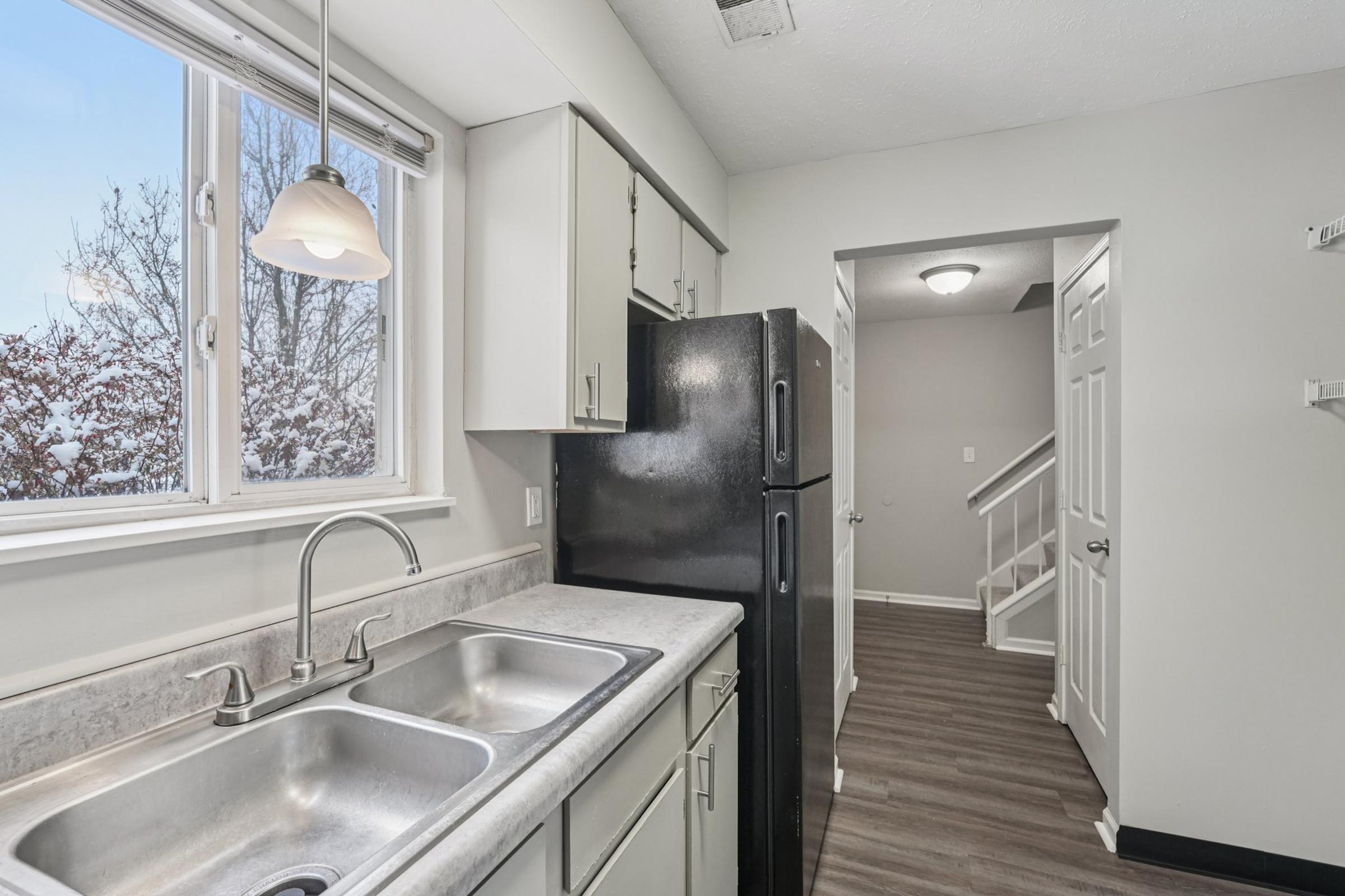 A modern kitchen with a double sink and a black refrigerator. There is a window above the sink allowing natural light to enter, and a glimpse of a hallway leading to stairs can be seen in the background. The walls are painted in a light color, and the flooring is made of laminate wood.
