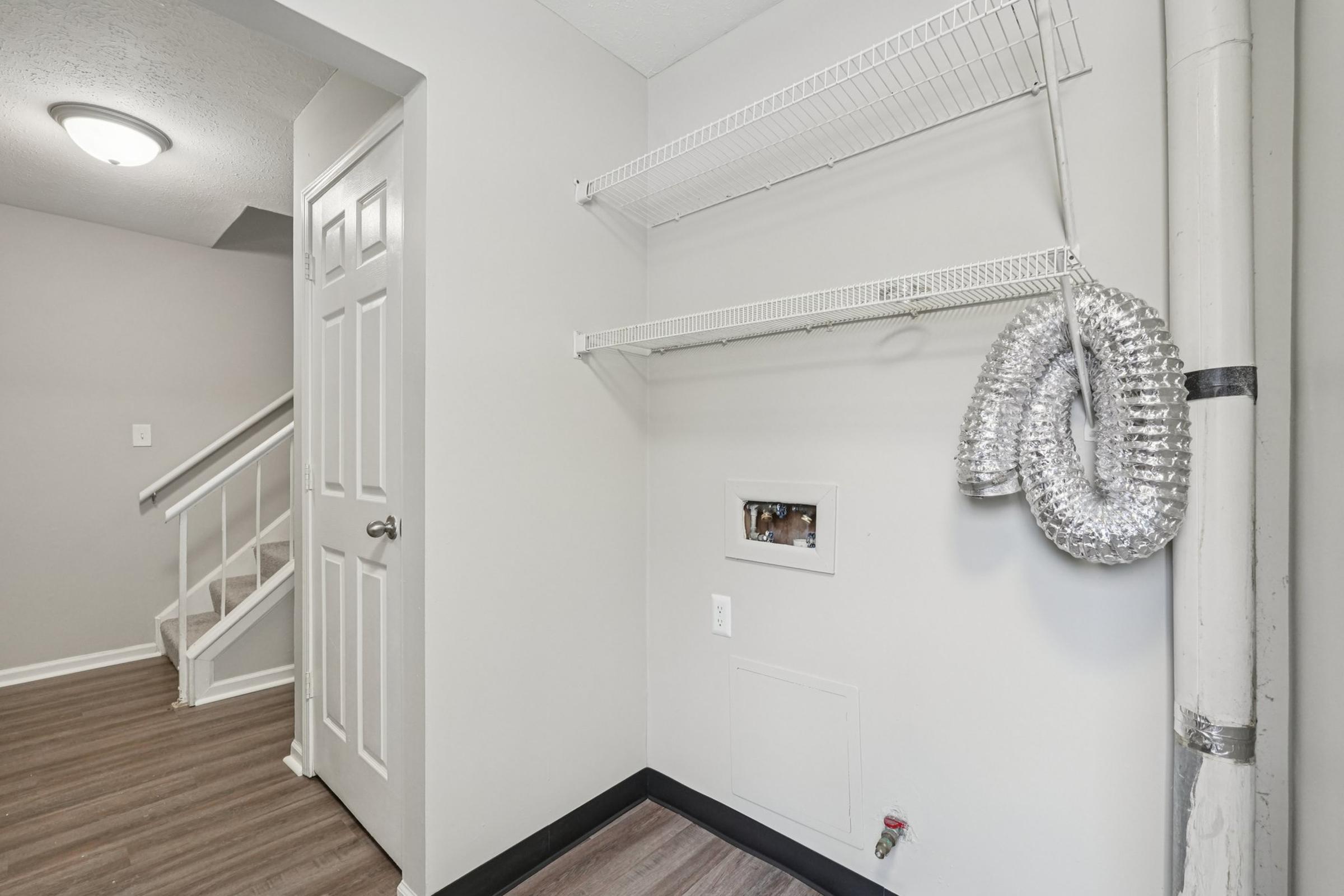 A small, empty laundry area featuring a shelf with wire racks on the wall, a vent hose hanging, and a wall-mounted electrical outlet. Stairs are visible in the background, leading to another level. The walls are painted light gray, and the flooring is wooden laminate.