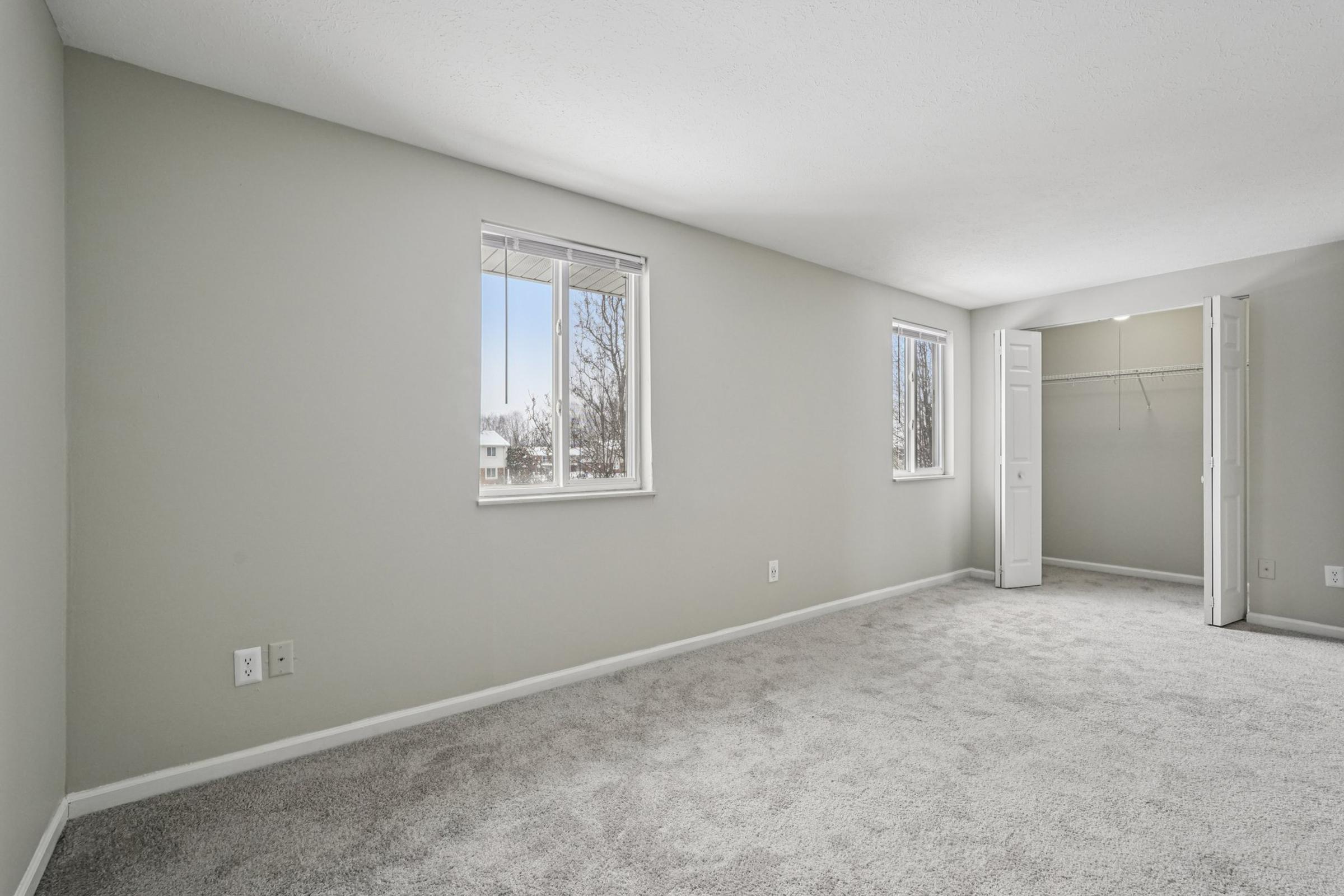 A spacious, empty room with light gray walls and plush carpet. Two windows allow natural light to fill the space, creating a bright atmosphere. A closet with open doors is visible, and the overall design offers a modern, minimalist aesthetic.
