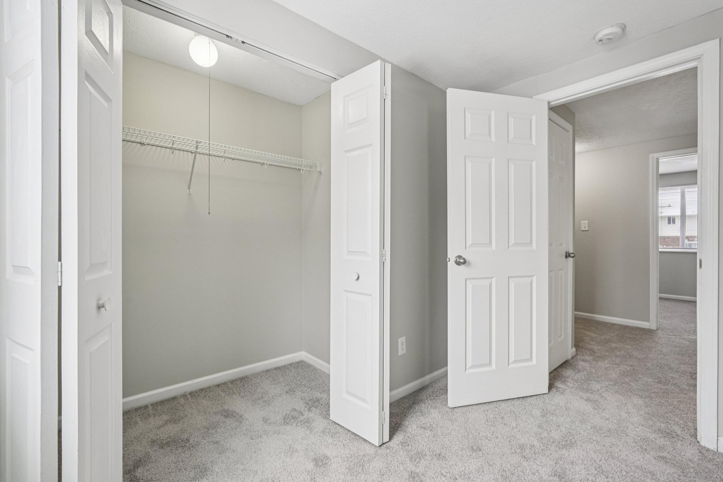 A well-lit, empty bedroom with light gray walls and plush carpet. Two open white doors reveal a spacious closet with a hanging rack. The room features a ceiling light fixture and a small window allowing natural light, creating a bright and airy atmosphere.