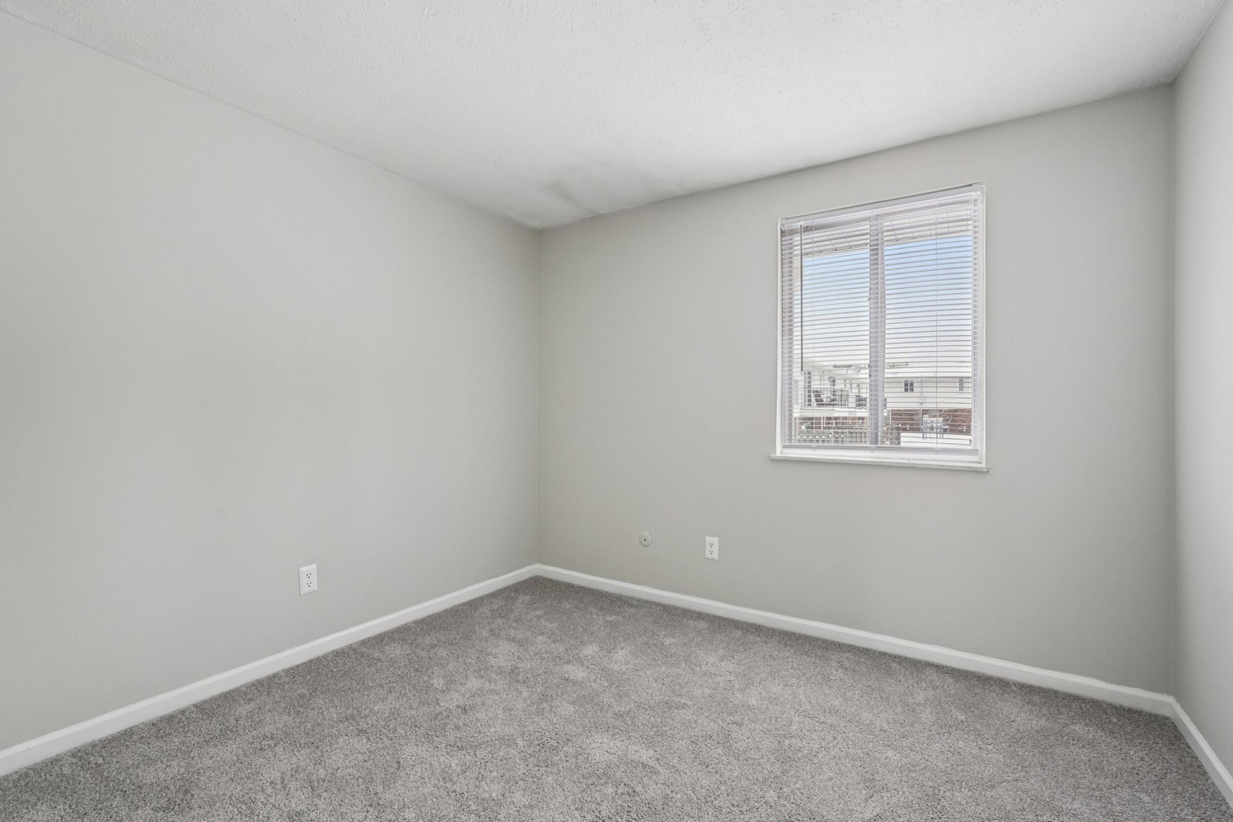 A vacant room with gray carpet and light gray walls. A single window on one wall lets in natural light, while the opposite wall features two electrical outlets. The space is empty and unfurnished, providing a clean and simple aesthetic.