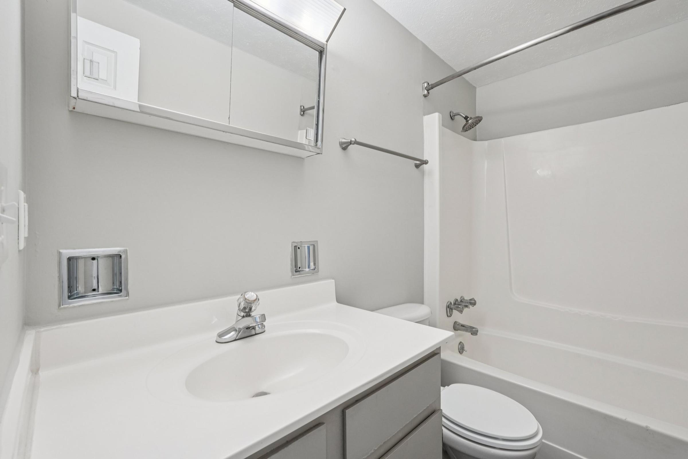 A clean and modern bathroom featuring a white sink with a single faucet, a shower/tub combination, a toilet, gray walls, and a mirror above the sink. The space is well-lit and organized with simple fixtures and limited decor.