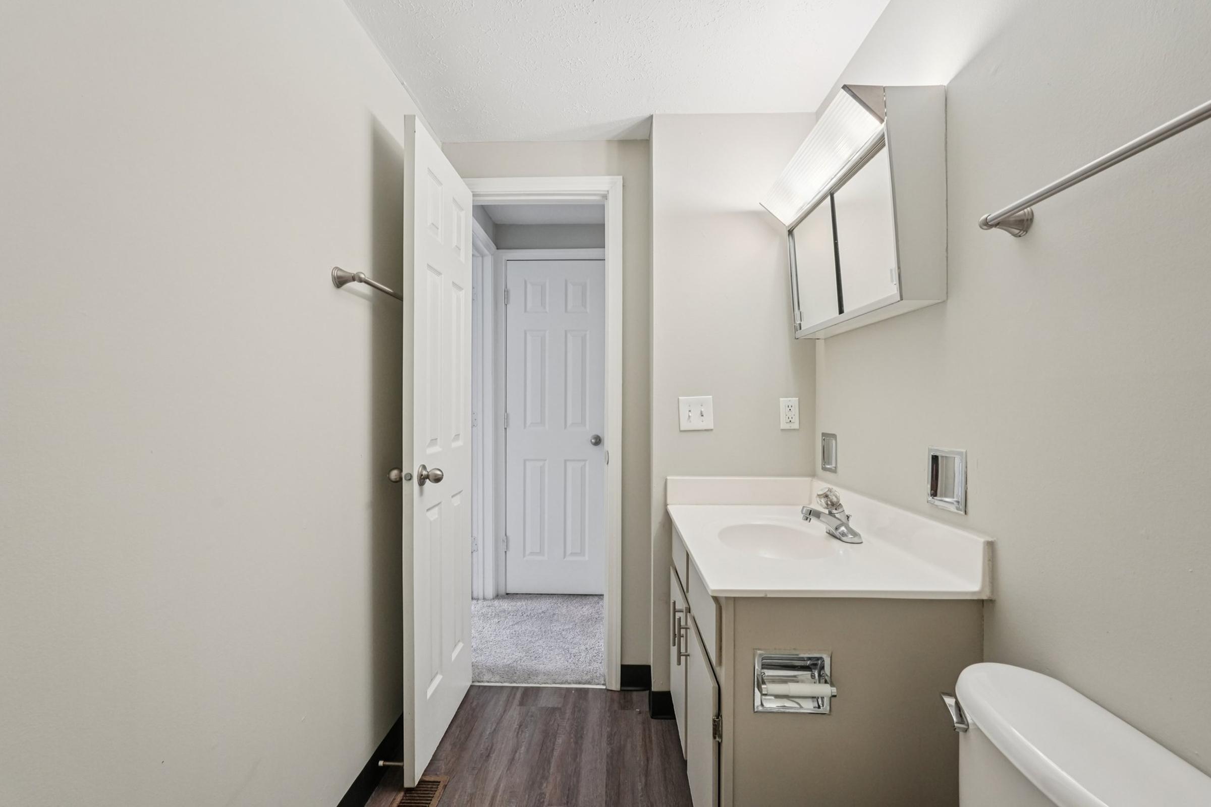 A clean, well-lit bathroom featuring a white vanity with a sink, a large mirror above, and gray walls. The flooring is dark, and a white toilet is visible. A partially opened door leads to another room, and a towel rack is mounted on the wall.