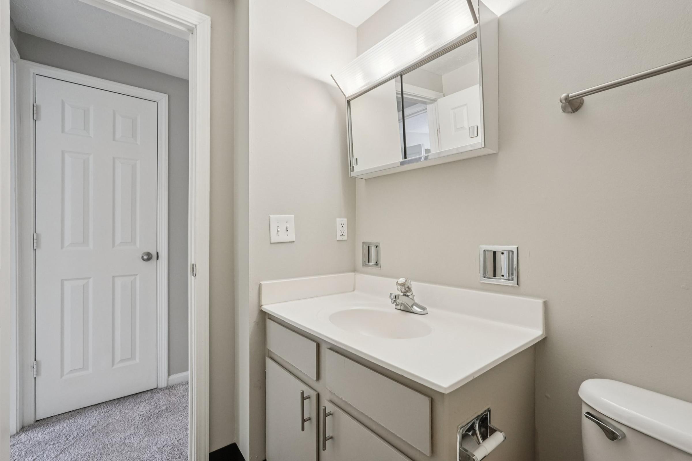 A small, well-lit bathroom featuring a white countertop with a sink, a mirror above it, and light-colored walls. To the right, there's a door leading to another room, and to the left, a towel rack is mounted on the wall. The floor is carpeted, creating a cozy feel.