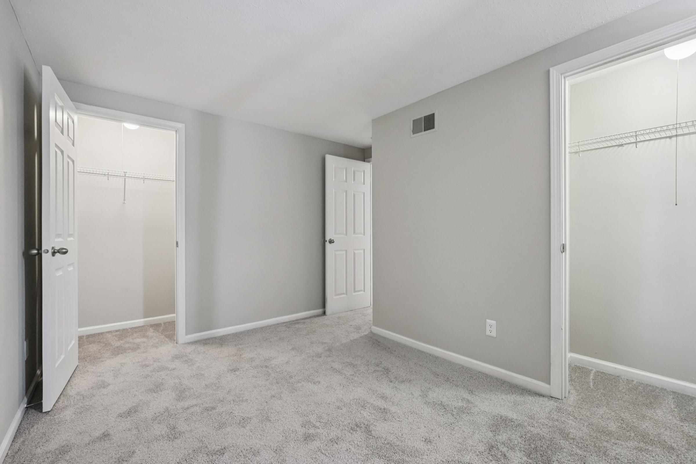 A spacious, neutral-colored bedroom featuring gray walls and carpet. Two doors lead to potential closets, and there's a well-lit entrance. The room is empty, creating a simple and clean look, ideal for personal decoration and styling.