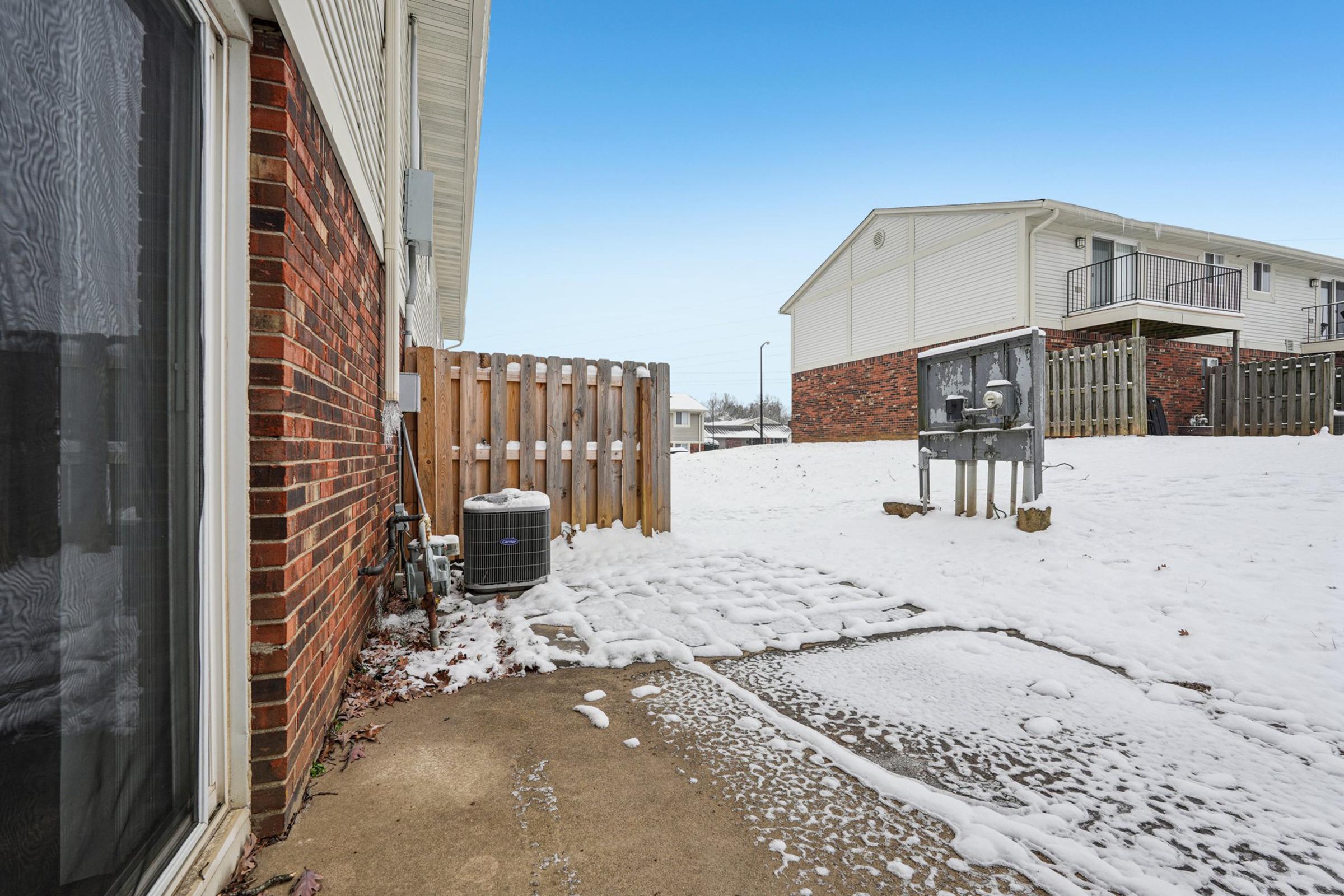 A snowy outdoor scene showing a paved area next to a building, with a wooden fence in the background. An air conditioning unit is visible near the wall, and a utility box or meter is seen nearby. The ground is partially covered in snow, and the sky is clear and blue.