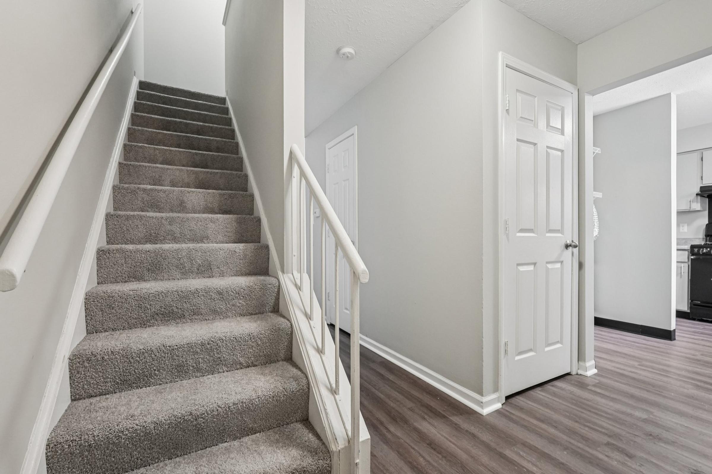 A well-lit hallway featuring a staircase with carpeted steps on the left, leading to an upper level. The walls are painted light gray, and there is a door on the right leading to another room. The flooring is a dark wood laminate, creating a modern and clean aesthetic.