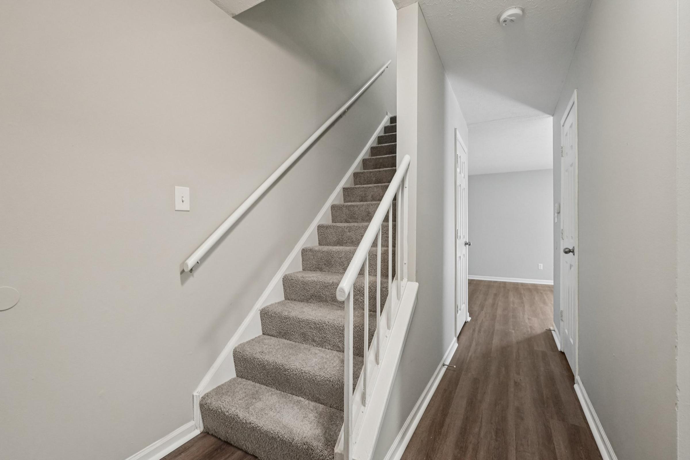 A well-lit interior hallway featuring a carpeted staircase to the left. The walls are painted in a light gray color, and there is a door on the right leading to another room. The flooring is a warm-toned laminate, creating a cozy atmosphere in the space.