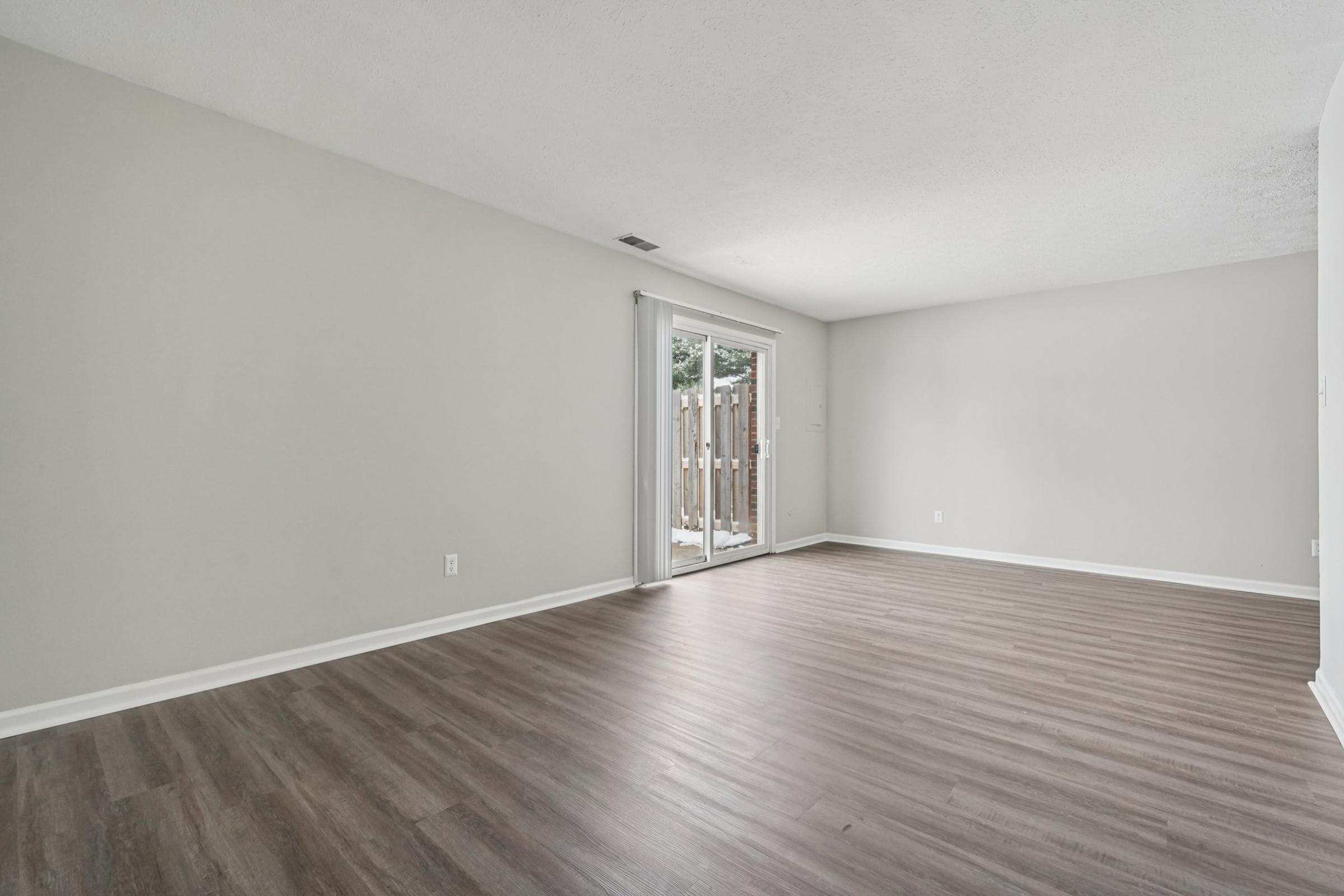 Spacious, empty room featuring light gray walls and hardwood-style flooring. A sliding glass door opens to a wooden-fenced outdoor area, allowing natural light to flow in. The space is uncluttered and ready for personalization.