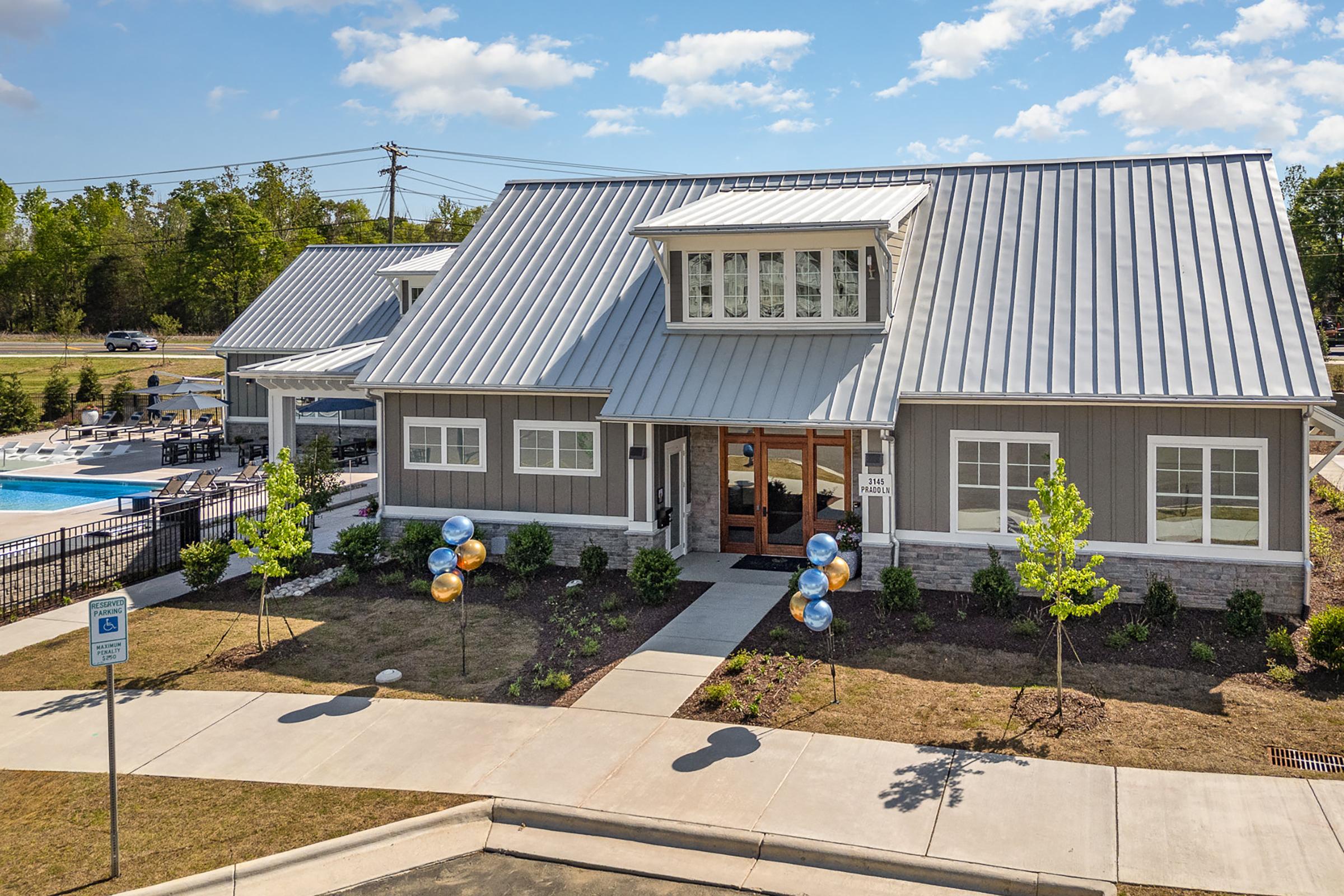 A modern clubhouse with a metal roof and large front windows, surrounded by landscaped greenery. In the foreground, a sidewalk leads to the entrance, and colorful balloons in blue and gold are positioned near the entrance. A swimming pool can be seen in the background.