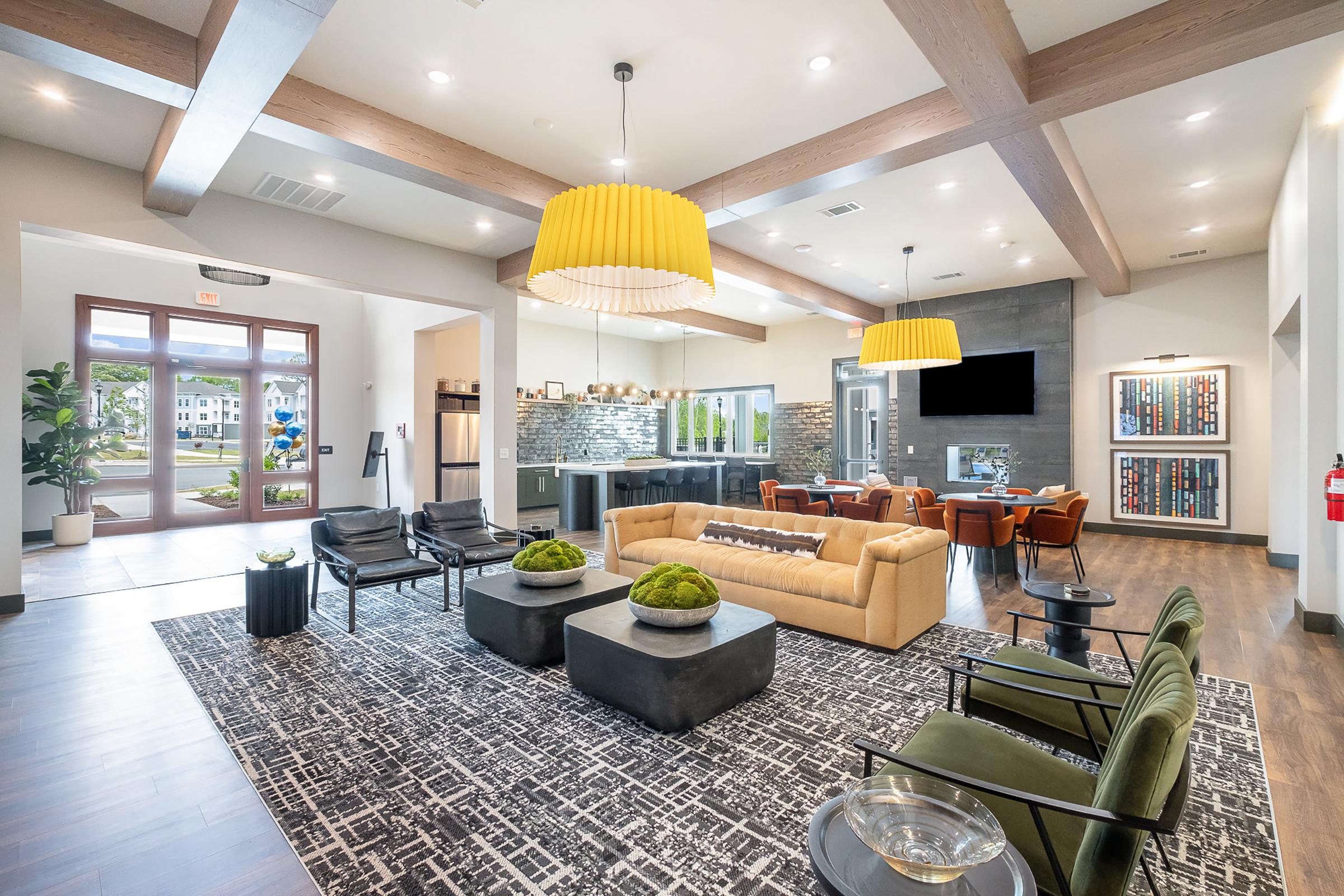 A modern lounge area featuring a stylish sofa and armchairs, large windows allowing natural light, contemporary pendant lights, and a decorative rug. The space includes a kitchenette and is accented with greenery, creating a welcoming atmosphere for socializing and relaxation.