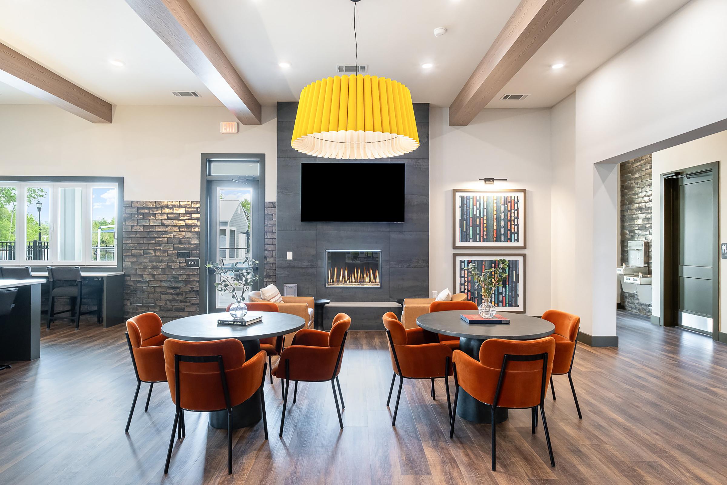 Modern interior of a spacious lounge area featuring several round tables with orange chairs, a large yellow pendant light overhead, and a stone wall with a fireplace. Large windows let in natural light, and decorative artwork adorns the walls, creating a warm and inviting atmosphere.