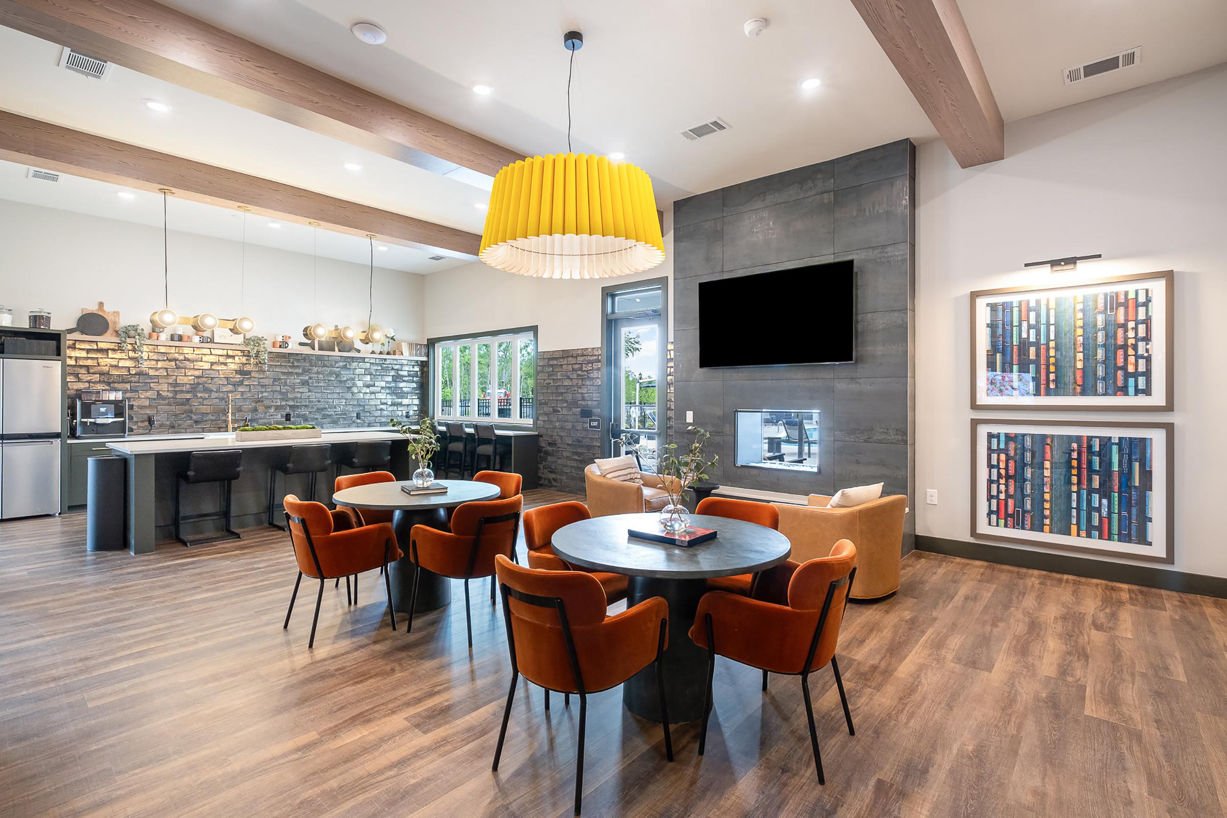 A modern communal space featuring stylish furnishings. The room has a high ceiling with wooden beams, a large decorative yellow pendant light, and a mix of seating options including round tables with orange chairs. A sleek kitchen area with bar seating is visible, along with a wall-mounted TV and framed artwork.