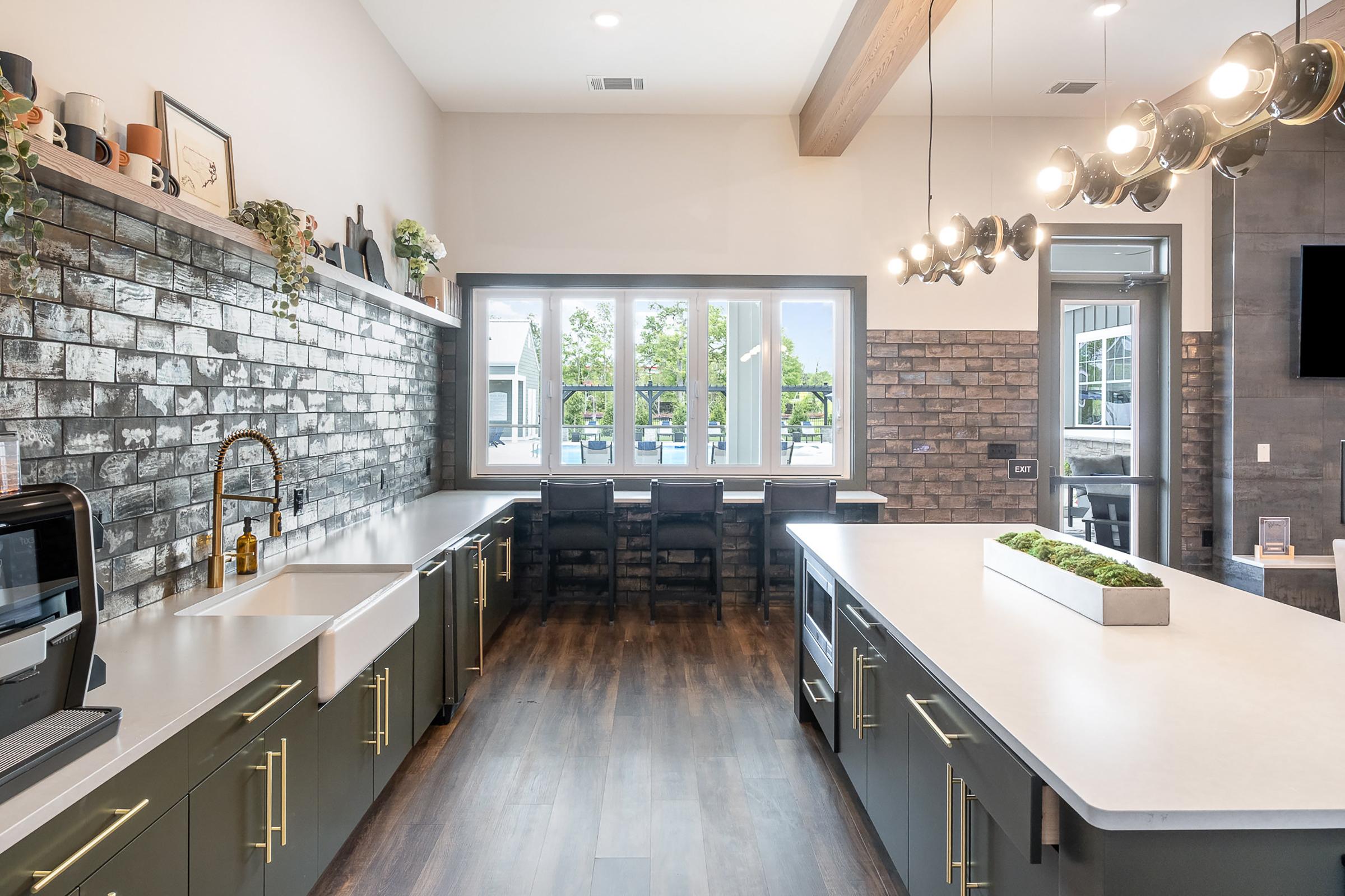 Modern kitchen featuring a spacious layout with dark cabinetry and gold hardware. The backsplash is made of textured gray tiles. A large window lets in natural light, illuminating the countertop and fixtures. There is a dining area with chairs visible through the window, creating an inviting atmosphere.