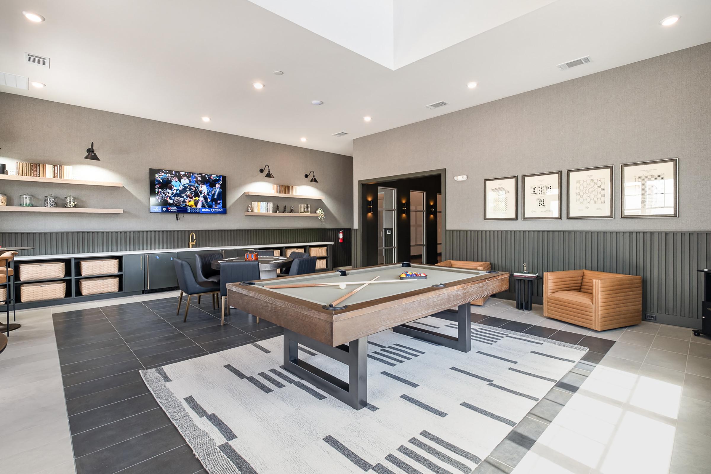 A modern game room featuring a pool table in the center, a large TV mounted on the wall displaying a sports event, stylish seating areas with a leather chair and black chairs, and decorative shelves with storage baskets. The walls are painted in neutral tones, creating an open and inviting atmosphere.